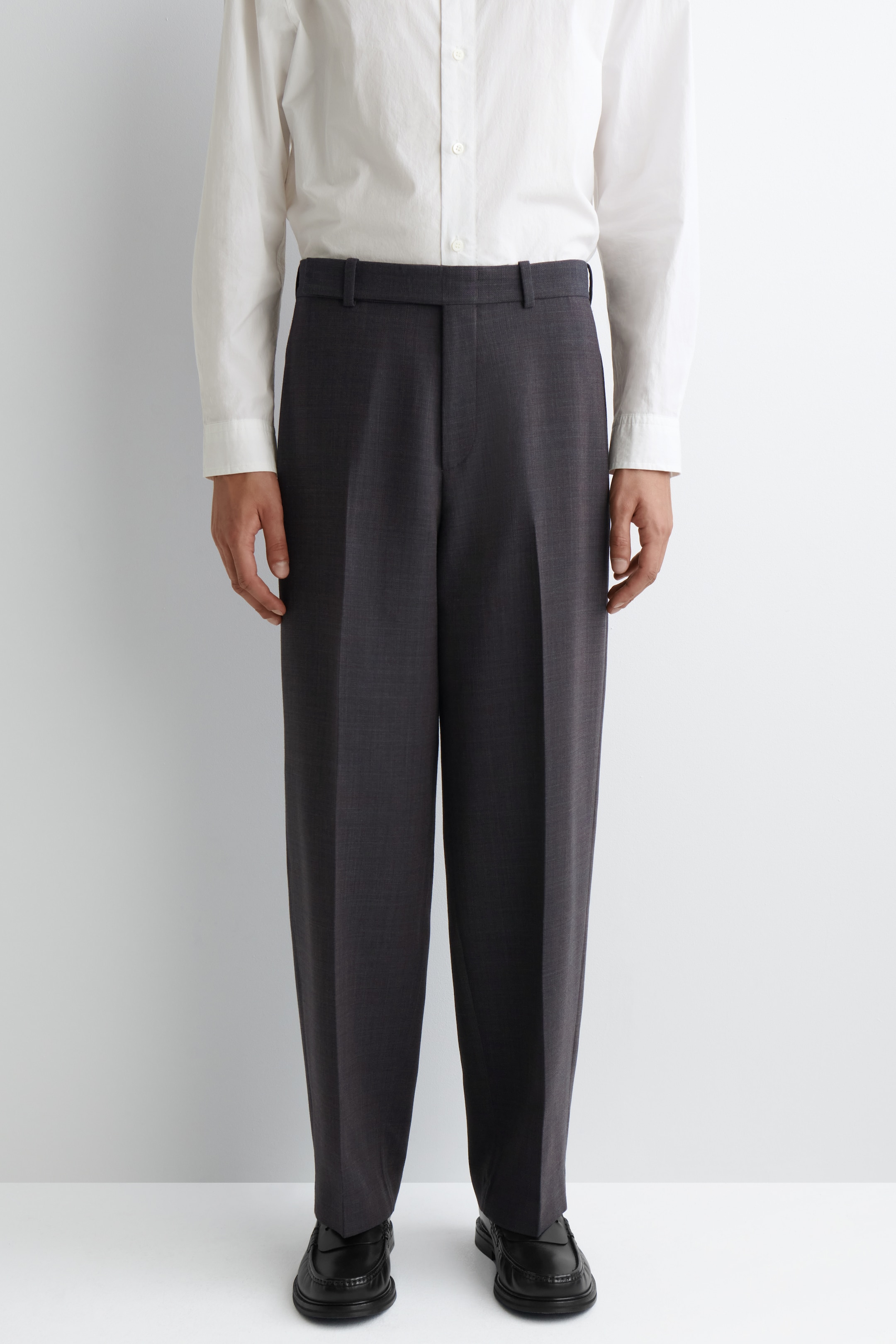 Agrandir l'image: Un pantalon large gris charbon foncé est porté, face à l'avant. Le pantalon taille haute présente des passants de ceinture visibles et des plis avant marqués, tombant librement sur des mocassins noirs.