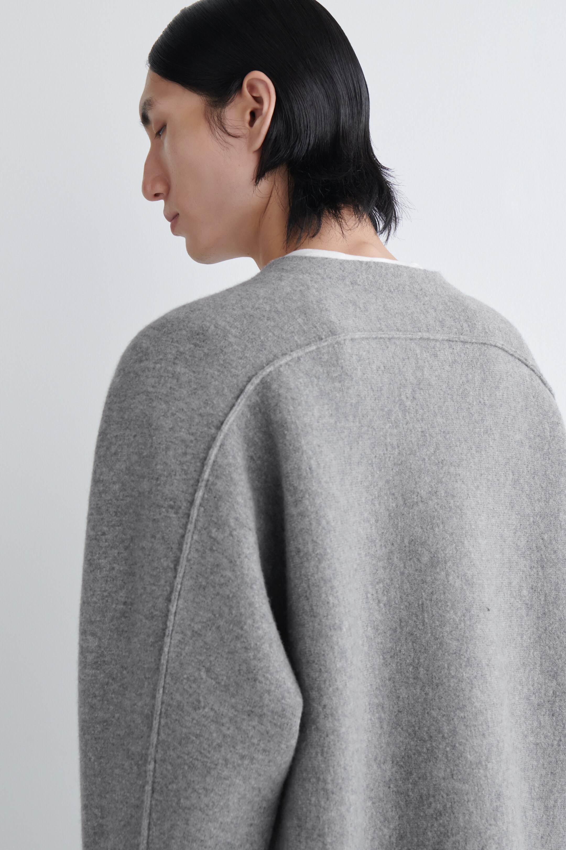 Agrandir l'image: Un pull gris est présenté de dos et de côté, montrant sa coupe décontractée. Il présente un col rond sans col, avec un col de t-shirt blanc visible en dessous. Des coutures distinctes définissent un empiècement arrière sur le haut du dos et descendent en courbe sur les épaules.