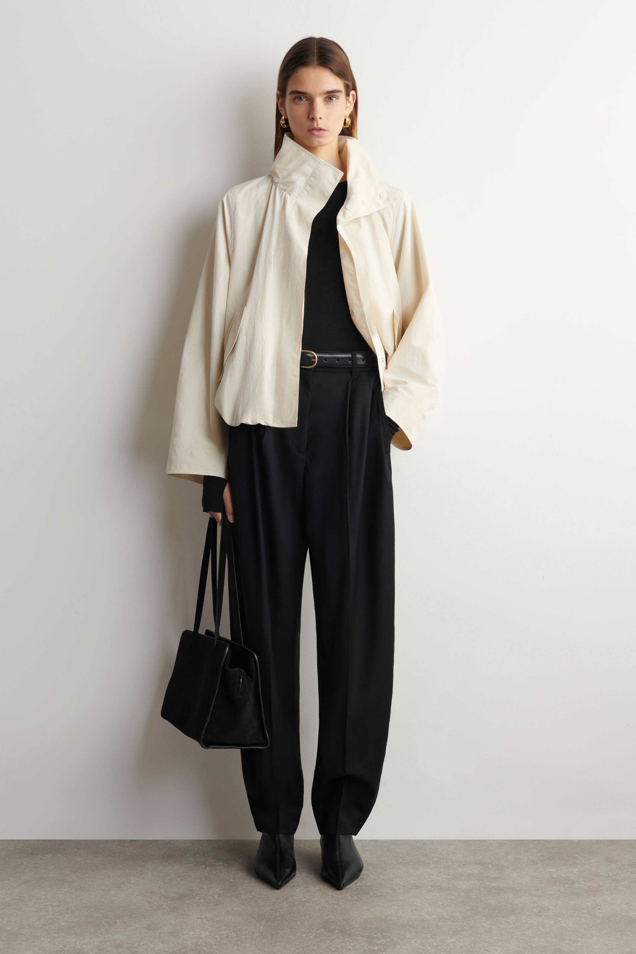 Agrandir l'image: Une veste courte crème, avec un col montant et des manches larges, est présentée de face, superposée à un haut noir à col montant. Un pantalon noir à jambes larges et plissées, maintenu par une ceinture noire, est porté avec des bottines noires à bout pointu, et un sac cabas structuré noir est porté.