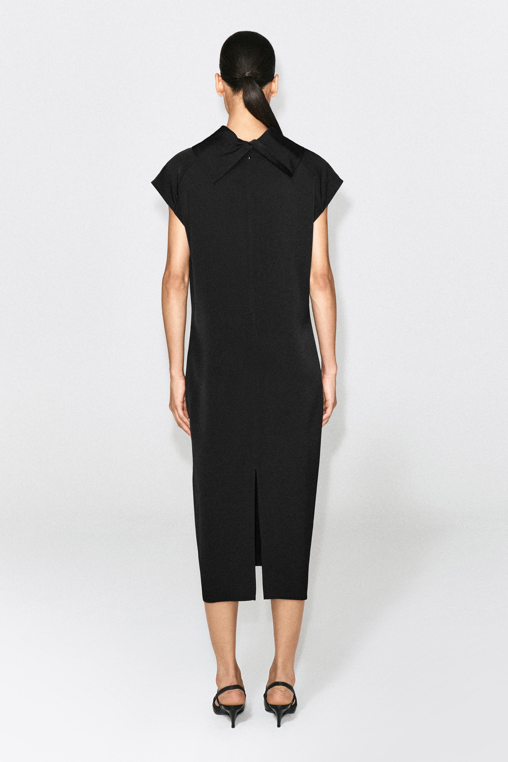 The black cowl-neck crepe midi dress, shown from the back, features a straight column silhouette with extended cap sleeves and a folded collar detail at the high neckline. The dress falls to a midi length and showcases a central back slit at the hem.