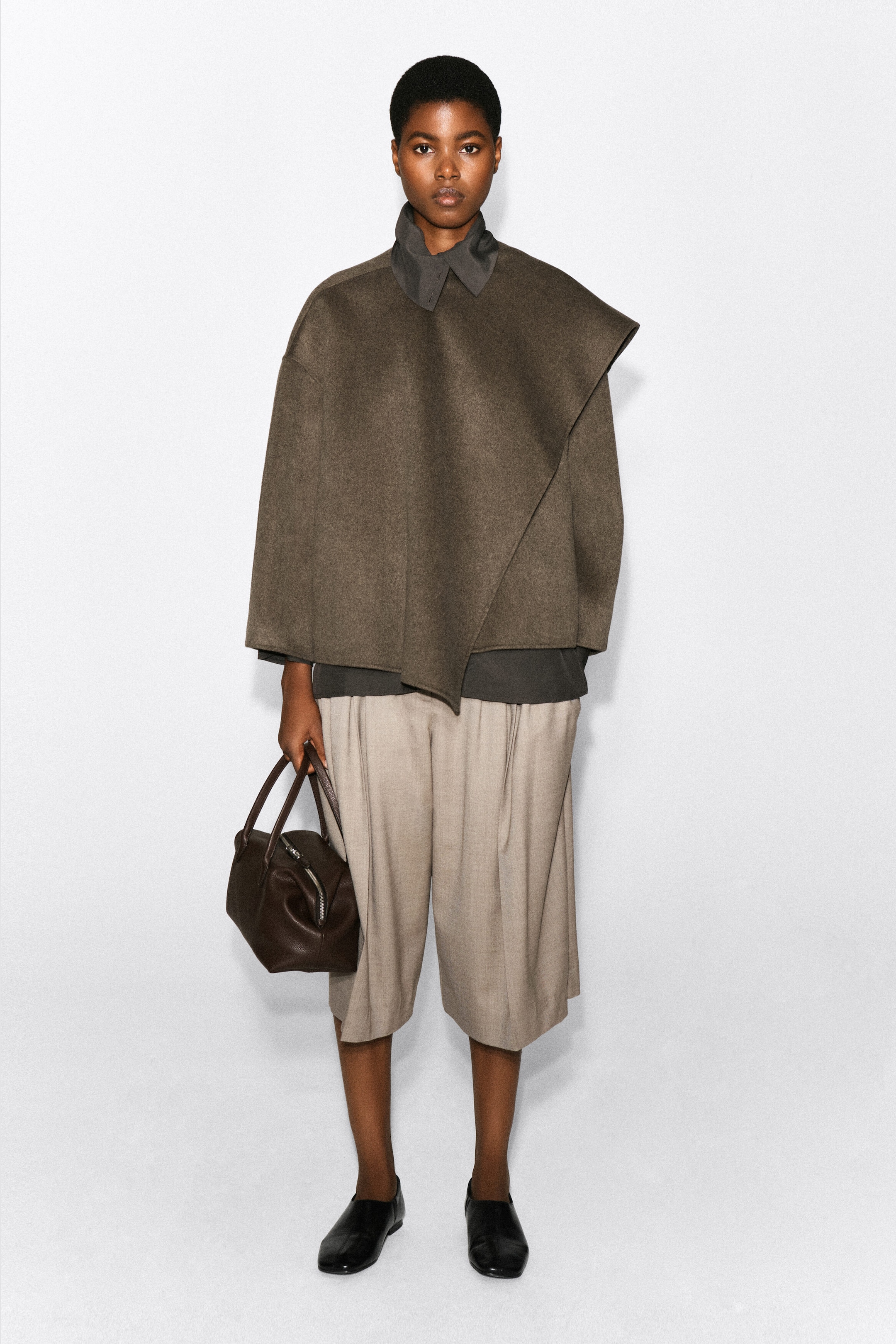 A double-faced wool-blend scarf jacket in a muted brown shade is being worn by the model, featuring an integrated, asymmetric draped front panel and relaxed wide sleeves. The jacket is layered over a dark collared shirt and styled with pleated beige culottes. A double-faced wool-blend scarf jacket in a muted brown shade is being worn by the model, featuring an integrated, asymmetric draped front panel and relaxed wide sleeves. The jacket is layered over a dark collared shirt and styled with pleated beige culottes.
