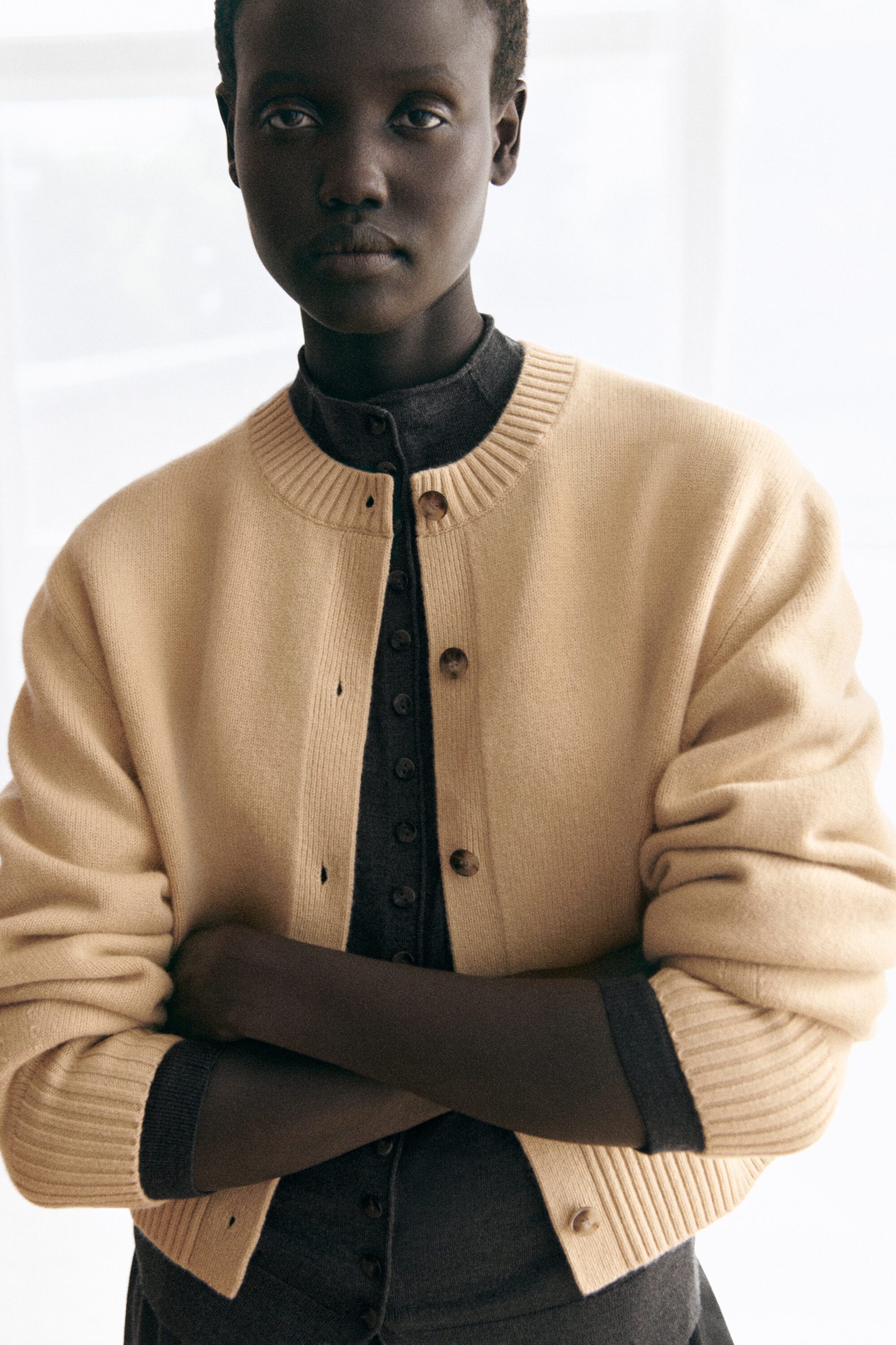 A beige merino wool cardigan featuring a ribbed crew neck and ribbed cuffs is worn open at the front, layered over a buttoned dark grey merino wool funnel-neck cardigan. A beige merino wool cardigan featuring a ribbed crew neck and ribbed cuffs is worn open at the front, layered over a buttoned dark grey merino wool funnel-neck cardigan.