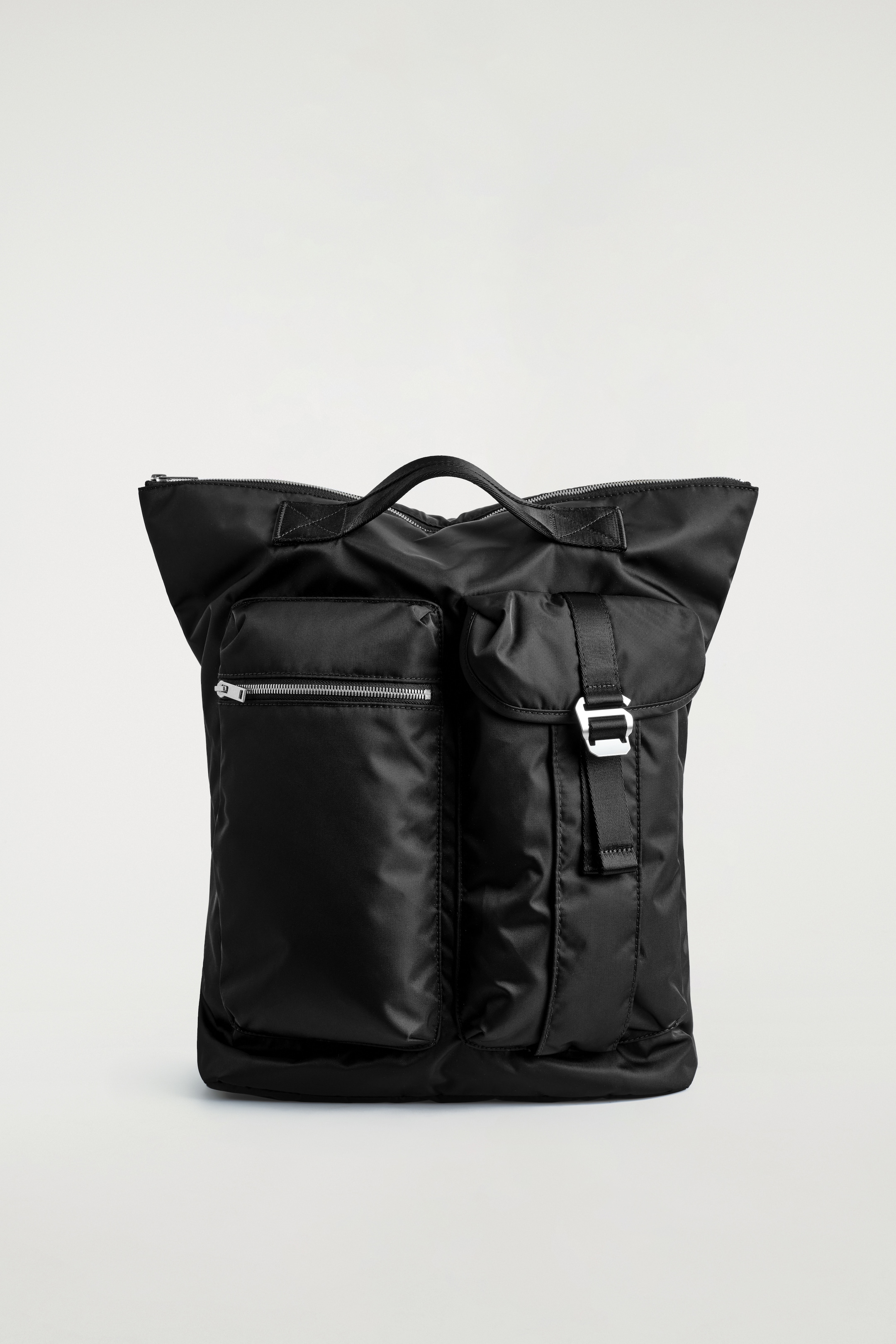View larger image: A black backpack is standing front-facing, featuring two top carry handles and a main zip closure. Its front is displaying two oversized pockets: a rectangular one with a silver-tone zipper on the left, and a larger flapped pocket on the right secured by a black strap and silver-tone buckle.
