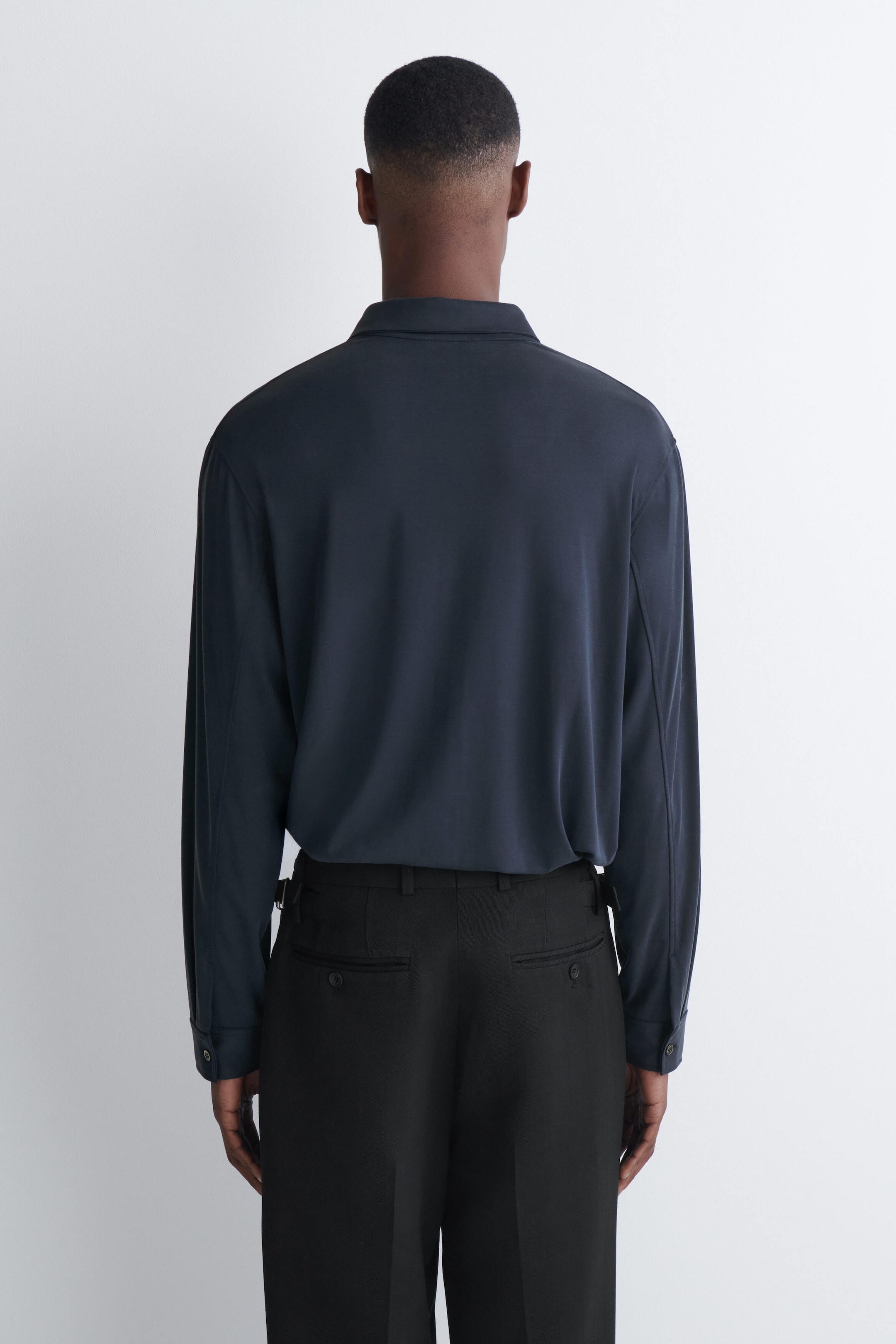 The fluid interlock jersey shirt in dark gray is worn by the model, shown from the back. The long-sleeved shirt features a soft collar and is tucked into tailored black trousers. The fluid interlock jersey shirt in dark gray is worn by the model, shown from the back. The long-sleeved shirt features a soft collar and is tucked into tailored black trousers.