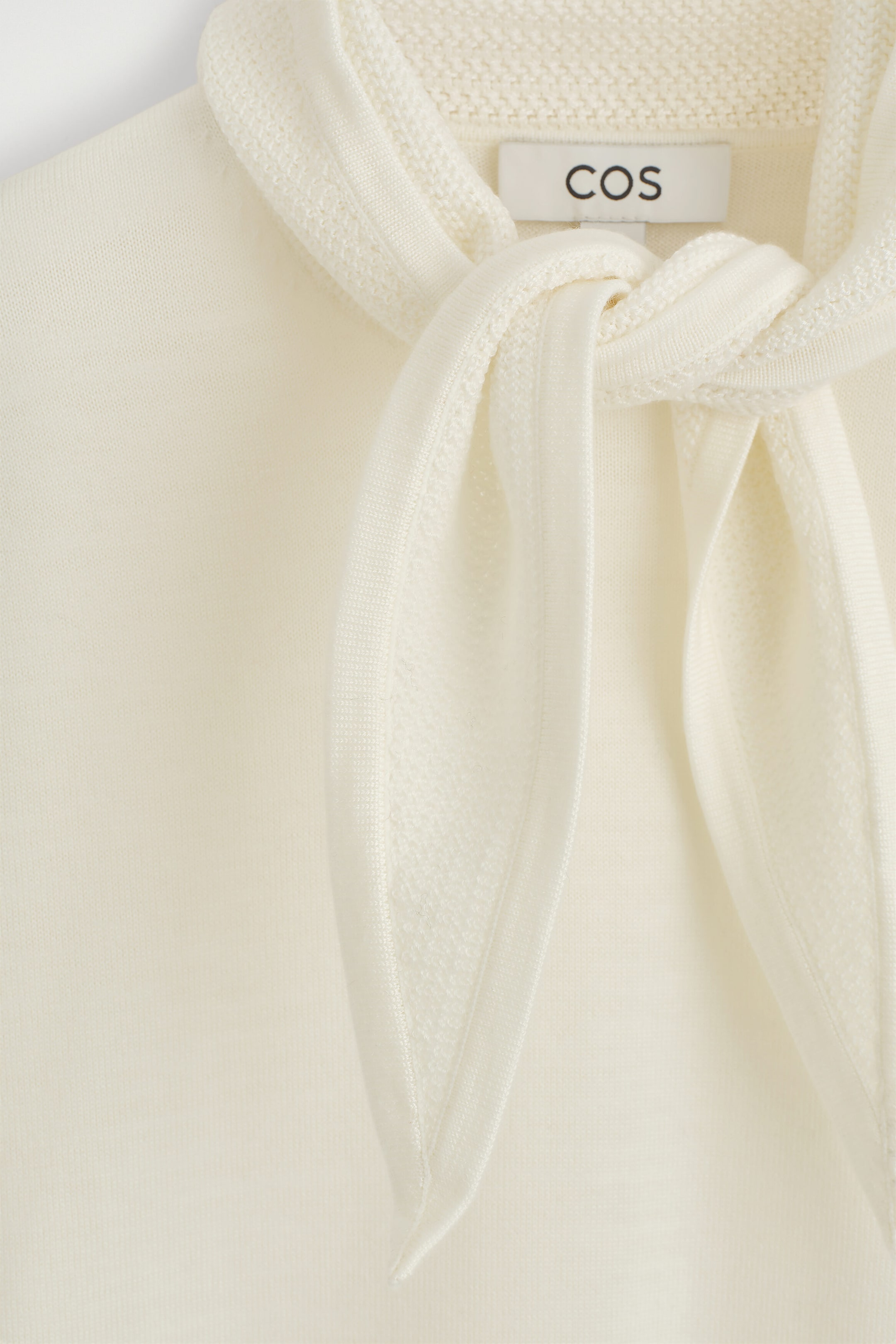 The cream-colored T-shirt in a merino wool blend with a scarf detail is shown in a close-up front view, highlighting its integrated collar tied in a loose knot at the neckline. The garment features a smooth, fine-knit body that contrasts with the textured knit of the draped scarf detail.