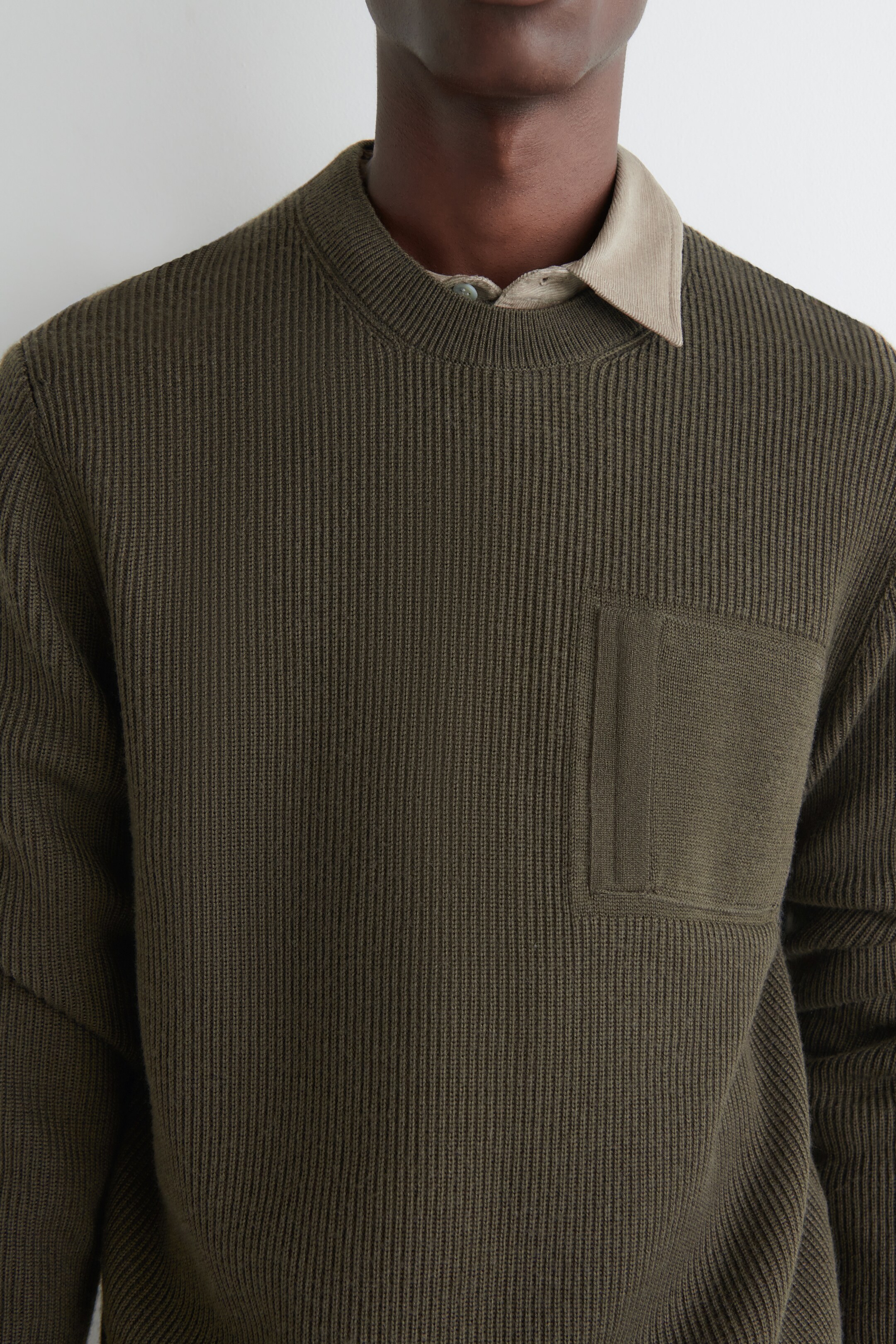 Agrandir l'image: Un pull vert olive est présenté, avec un tricot côtelé vertical et un col rond. Une poche plaquée avec un tricot plus lisse est visible sur la poitrine gauche. Une chemise à col clair est superposée en dessous, avec son col qui dépasse de l'encolure.