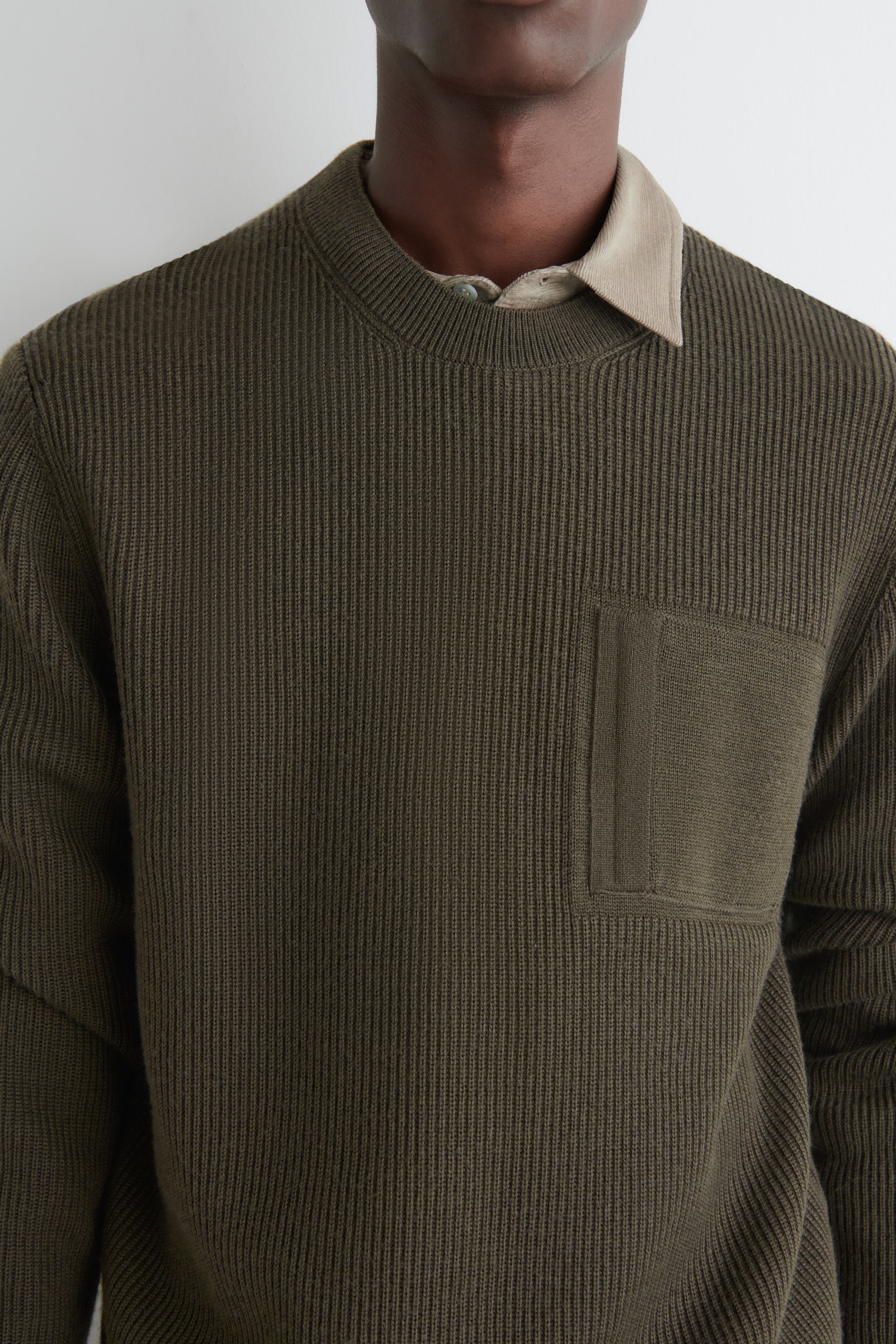 Grösseres Bild ansehen: Ein olivgrüner Pullover wird präsentiert, der ein vertikal geripptes Strickmuster und einen Rundhalsausschnitt aufweist. Eine aufgesetzte Tasche mit einem glatteren Strick ist auf der linken Brust sichtbar. Ein helles Hemd mit Kragen wird darunter getragen, wobei der Kragen aus dem Ausschnitt hervorschaut.
