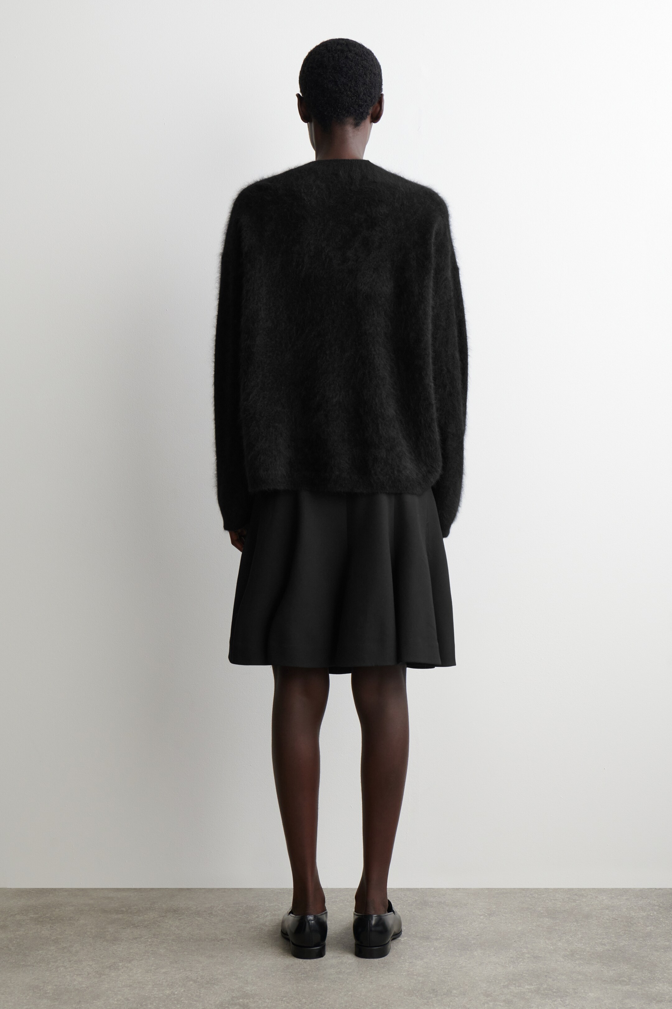 Se større billede: En overdimensioneret sort, fuzzy strikket sweater bæres, set bagfra, lagdelt over en sort, struktureret, udsvingende mininederdel. Sorte flade sko bæres også.