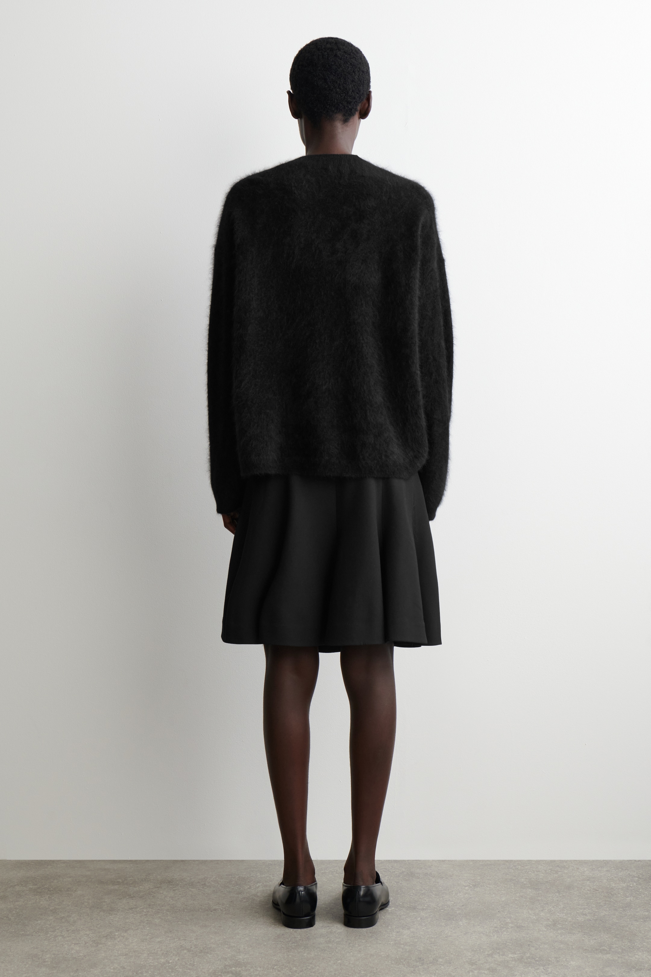 Grösseres Bild ansehen: Ein übergroßer schwarzer flauschiger Strickpullover wird getragen, von hinten gesehen, über einem schwarzen, strukturierten, ausgestellten Minirock geschichtet. Schwarze flache Schuhe werden ebenfalls getragen.