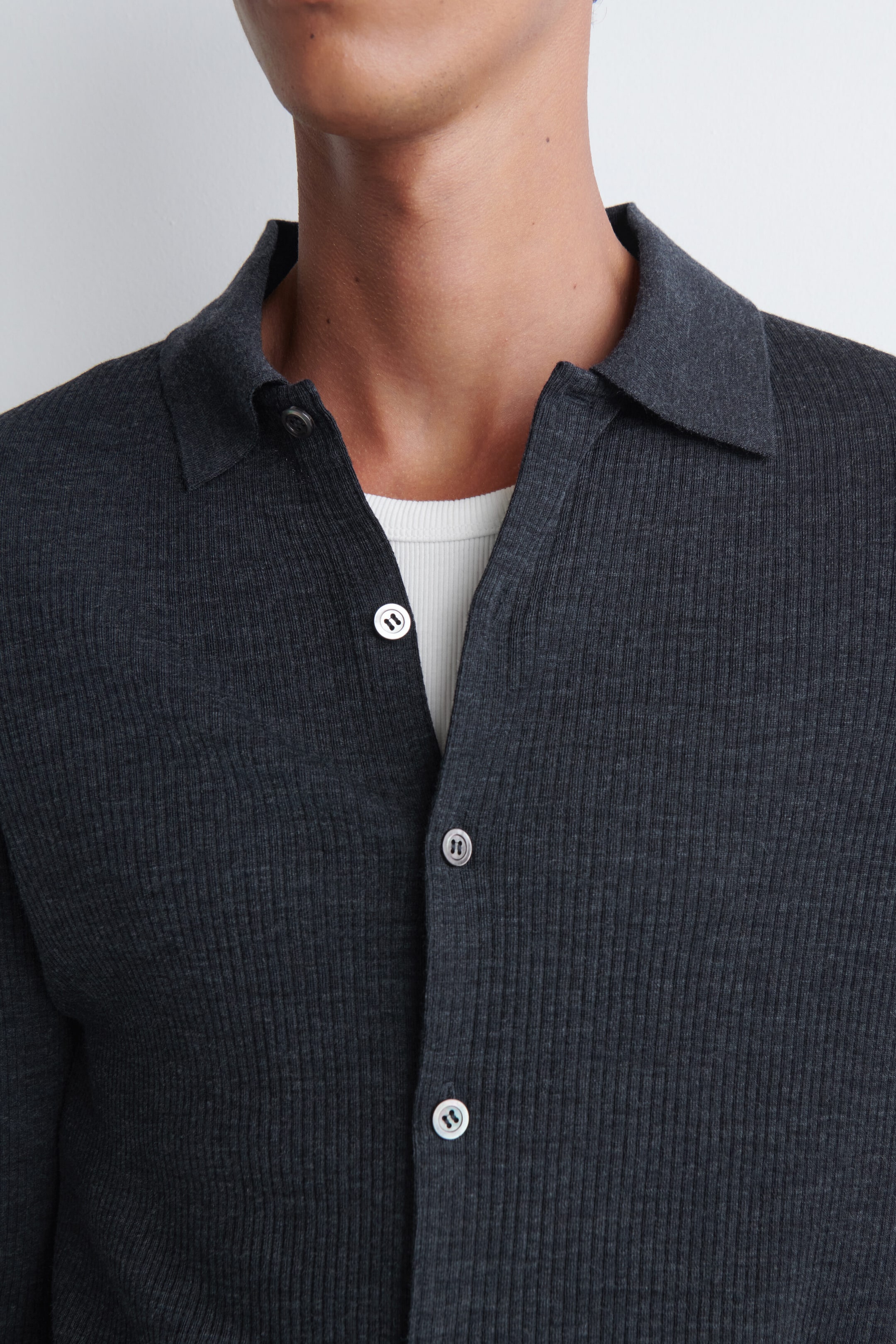 A dark grey ribbed knit button-up shirt featuring a classic collar and light grey buttons is being worn by the model. The shirt is styled open at the collar, revealing a layered white ribbed undershirt underneath.