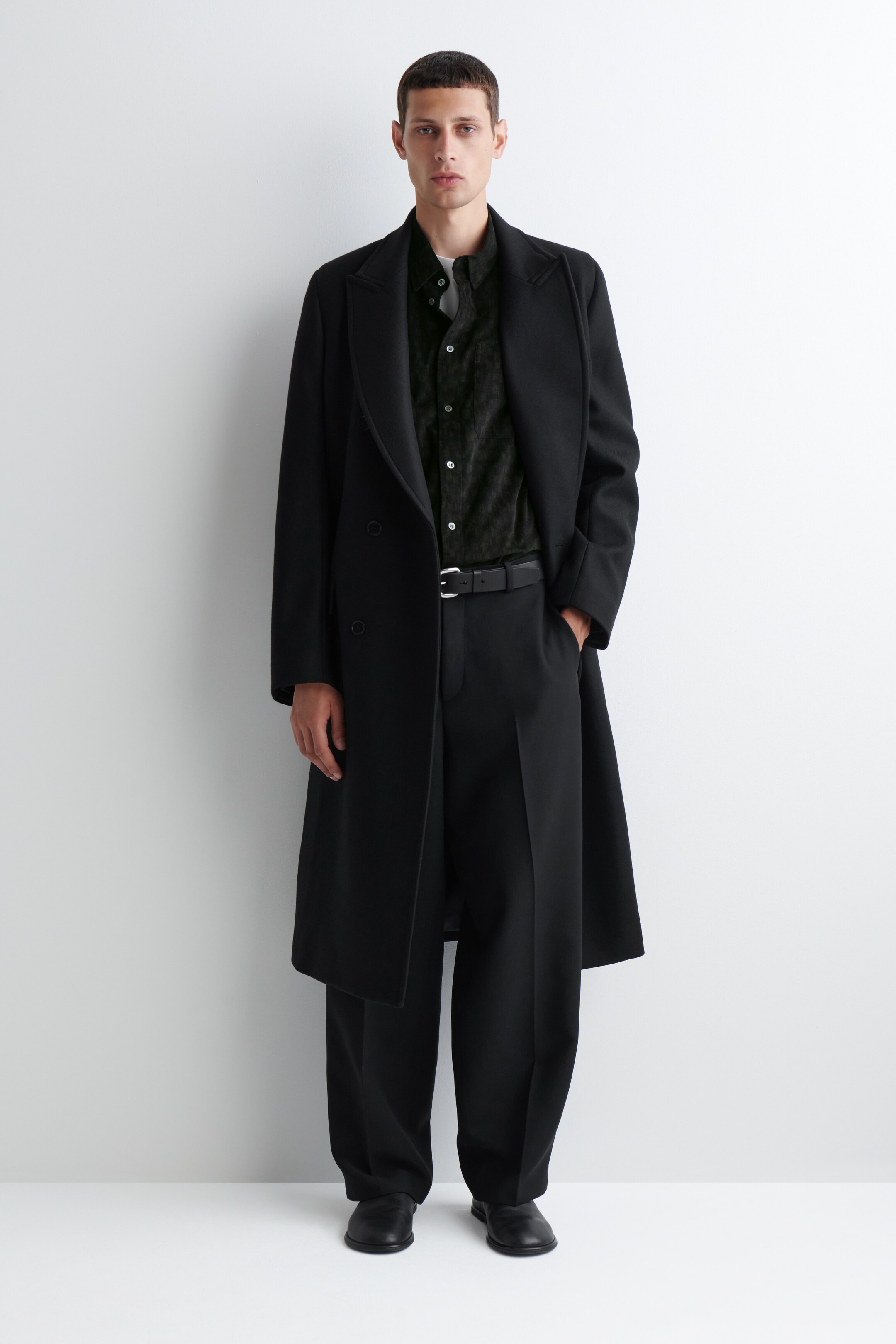 Agrandir l'image: La chemise en velours côtelé noir est présentée de face, avec une coupe décontractée et une fermeture à boutons avec des boutons de couleur claire, et est partiellement visible sous un long manteau noir.