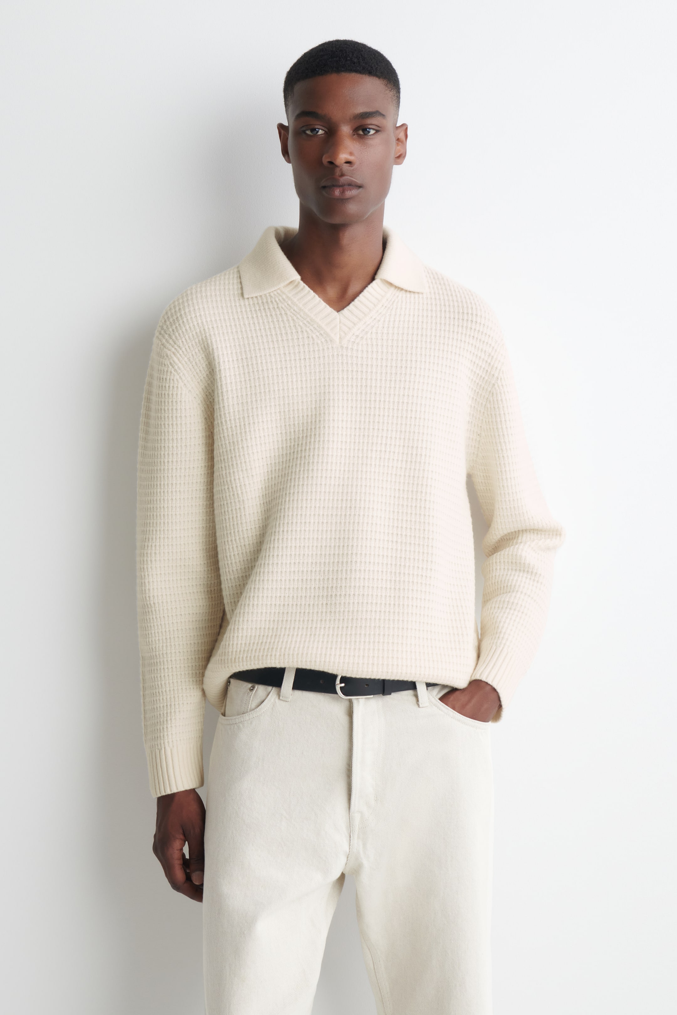 Visualizza immagine più grande: La modella indossa un maglione polo in lana a maglia waffle color crema, con colletto senza bottoni e scollo a V. Il maglione è infilato morbidamente in pantaloni chiari con una cintura nera.