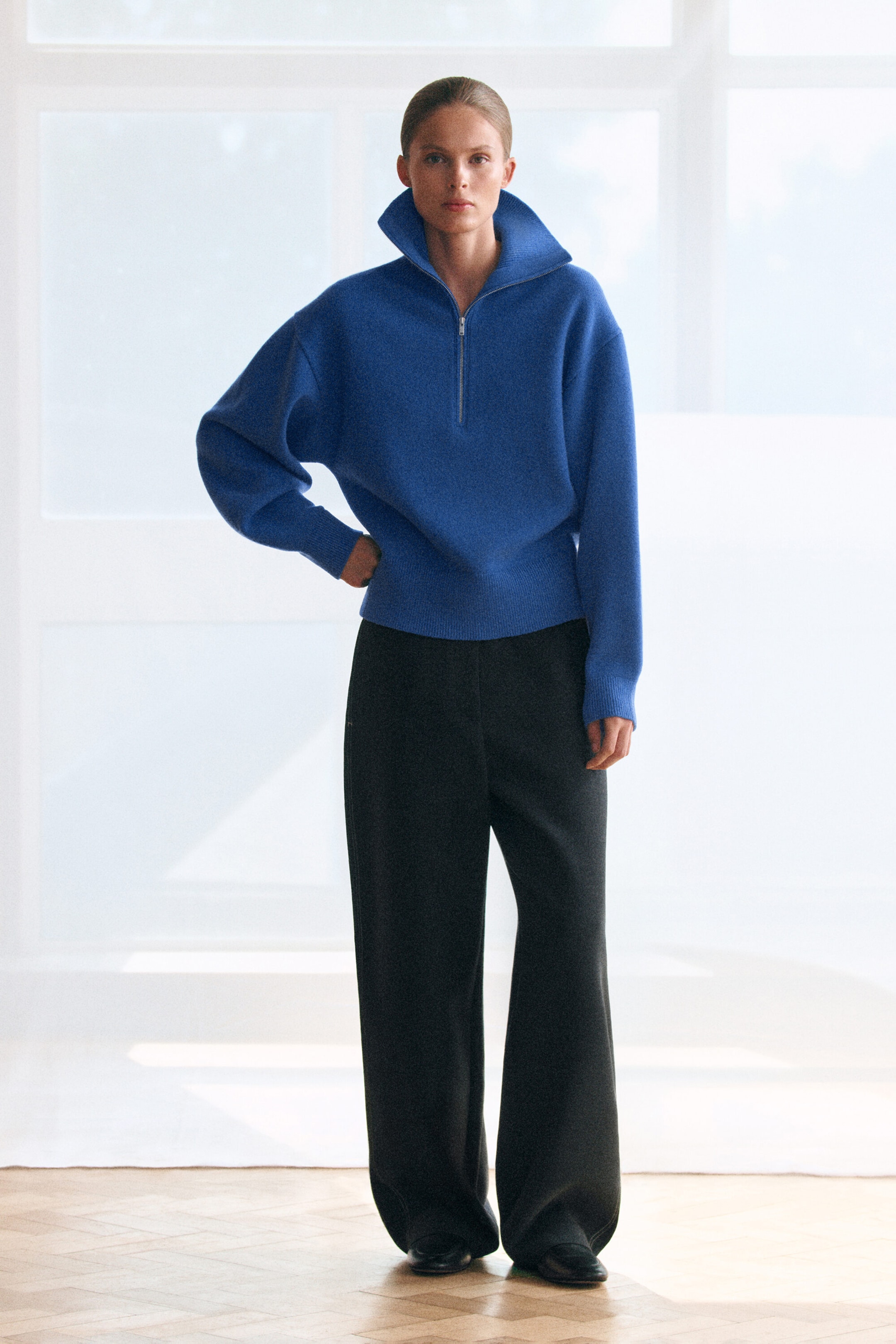 A royal blue knitted jumper is shown from the front, featuring a relaxed fit, sculptural sleeves with ribbed cuffs, and a silver-tone half-zip closure at the funnel neckline. The jumper is styled with wide-leg black trousers. A royal blue knitted jumper is shown from the front, featuring a relaxed fit, sculptural sleeves with ribbed cuffs, and a silver-tone half-zip closure at the funnel neckline. The jumper is styled with wide-leg black trousers.
