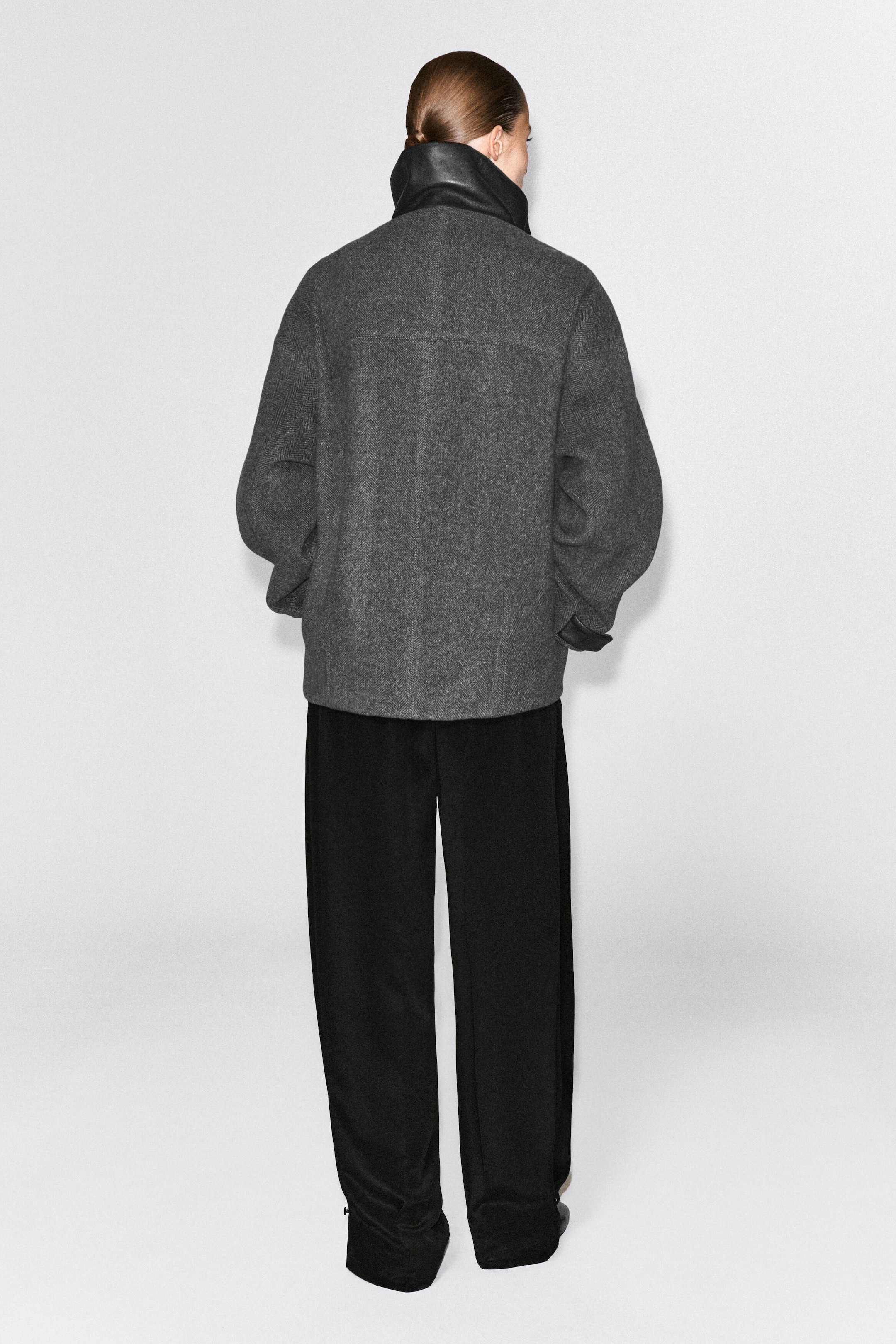 Ein graue Fischgrat-Jacke aus Wollmischung mit hohem schwarzem Leder-Stehkragen wird vom Model von hinten getragen. Die Oversize-Jacke ist mit schwarzen Wide-Leg-Hosen kombiniert und betont ihre lockere Passform sowie die dunklen Lederdetails an den Manschetten. Ein graue Fischgrat-Jacke aus Wollmischung mit hohem schwarzem Leder-Stehkragen wird vom Model von hinten getragen. Die Oversize-Jacke ist mit schwarzen Wide-Leg-Hosen kombiniert und betont ihre lockere Passform sowie die dunklen Lederdetails an den Manschetten.