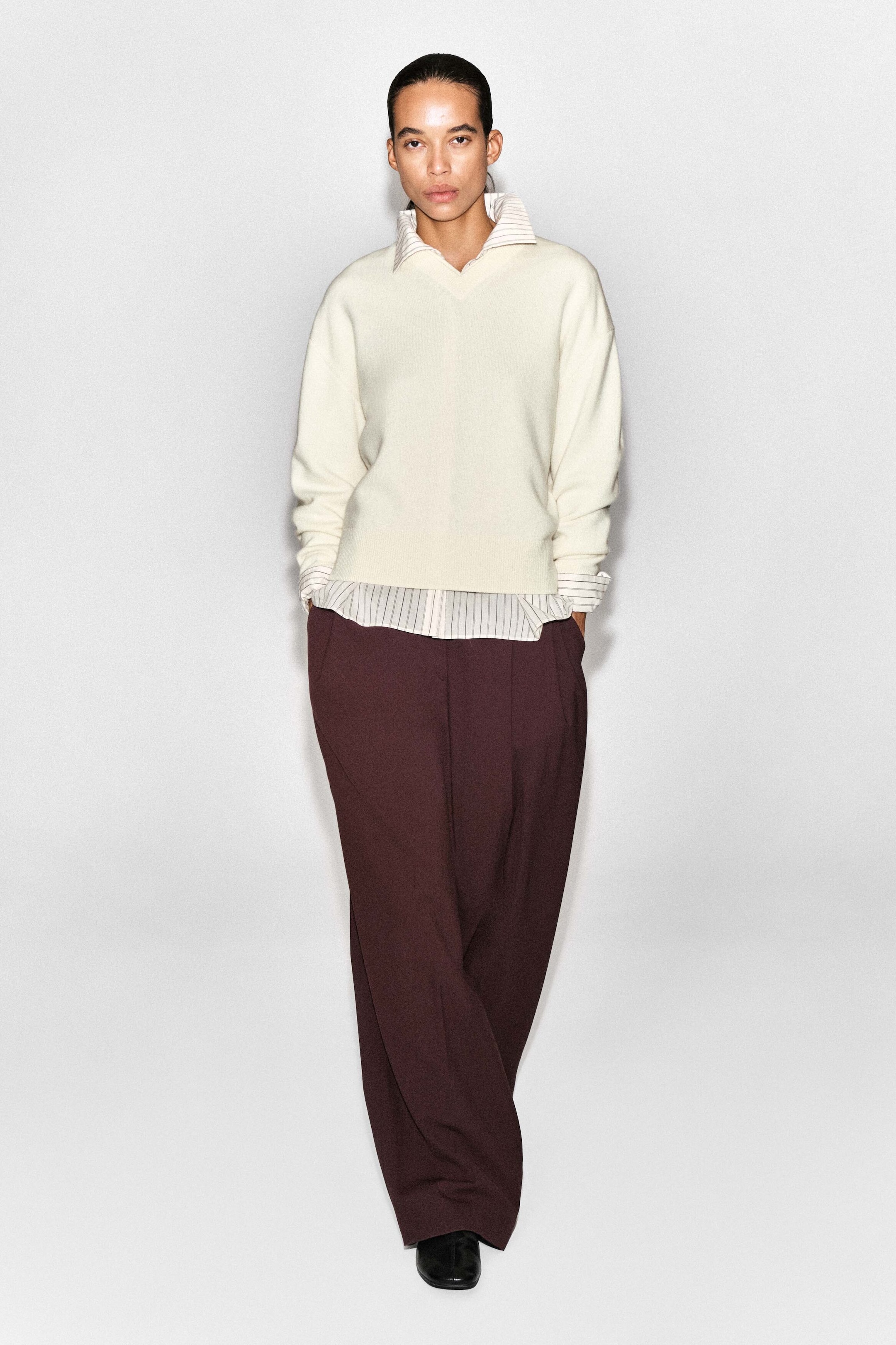 The merino wool layered V-neck jumper is shown from the front, featuring a relaxed fit, a V-neckline, and ribbed trims. It shows a striped collared shirt layer peeking out at the neckline, cuffs, and hem, and is styled with dark burgundy wide-leg trousers. The merino wool layered V-neck jumper is shown from the front, featuring a relaxed fit, a V-neckline, and ribbed trims. It shows a striped collared shirt layer peeking out at the neckline, cuffs, and hem, and is styled with dark burgundy wide-leg trousers.