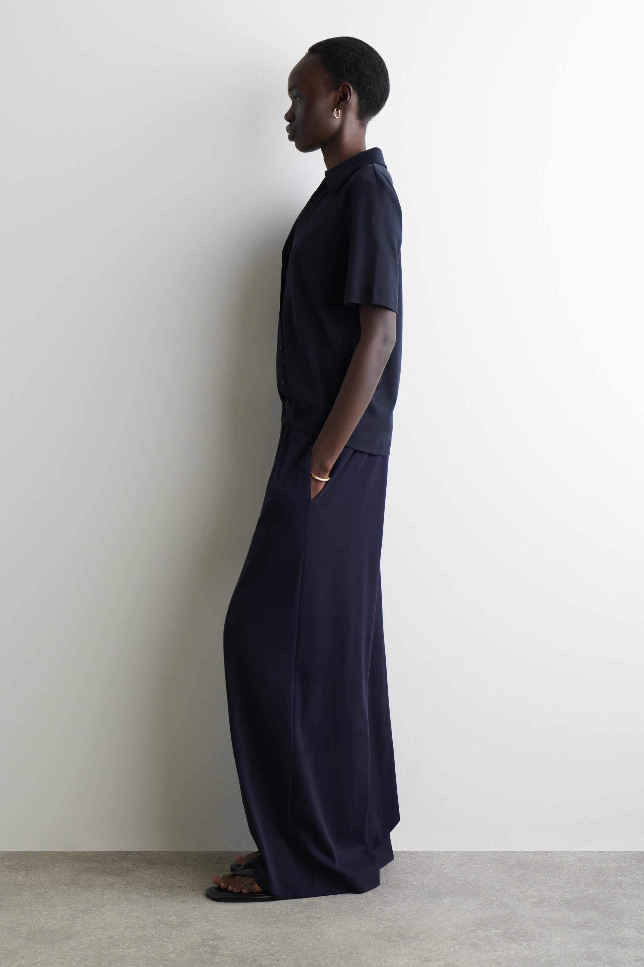 Agrandir l'image: Une chemise bleu marine à manches courtes avec col, dotée d'une fermeture à boutons sur le devant et d'une coupe décontractée, est portée non rentrée sur un pantalon bleu marine à jambes larges, stylisé avec des poches latérales et un design fluide et long, le tout présenté de profil.