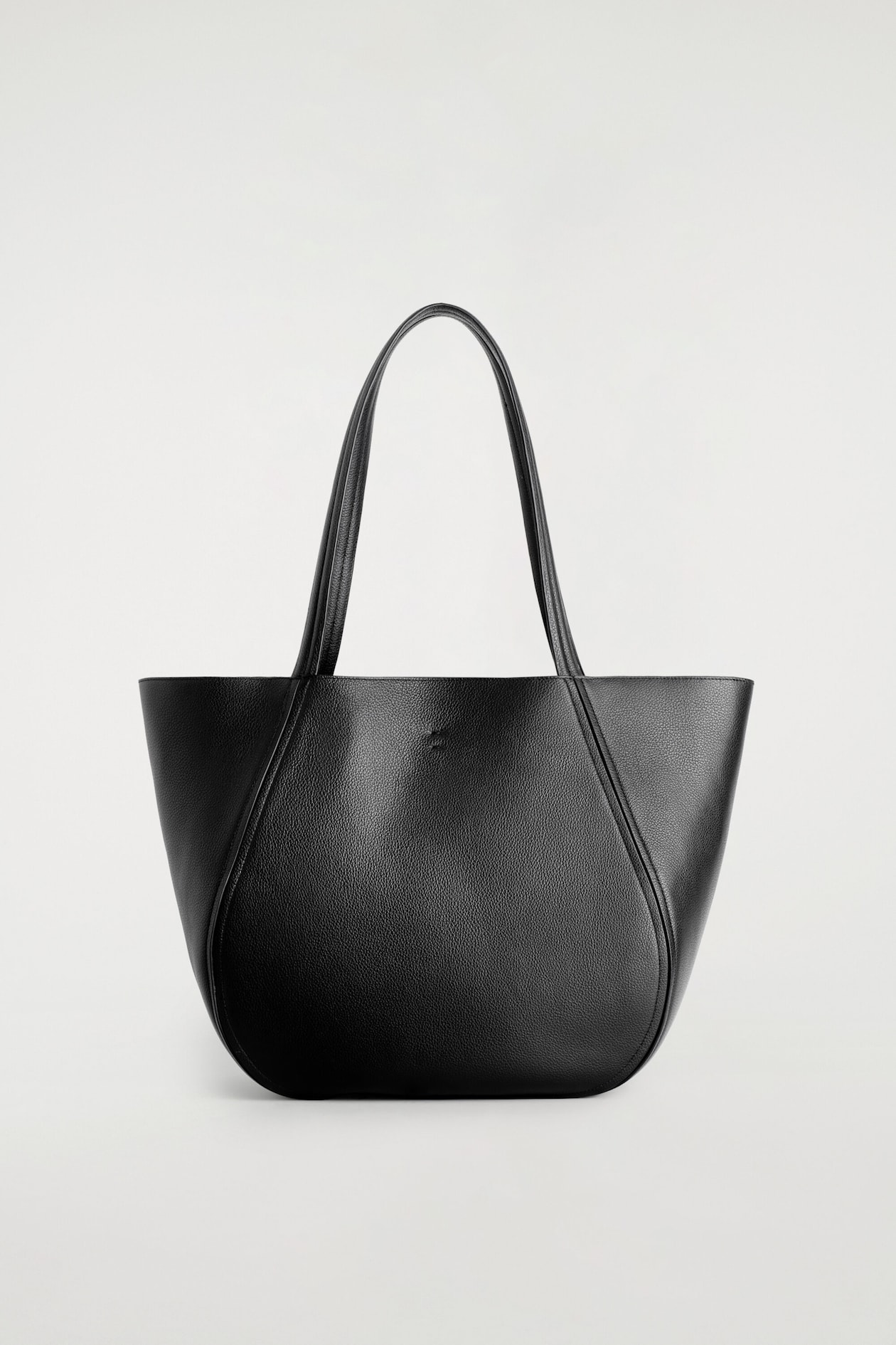 A black grained leather shopper bag is shown from the front, featuring a softly curved silhouette with pleated seams framing the center panel. Two matching top handles extend from the seams, highlighting the bag’s textured finish.
