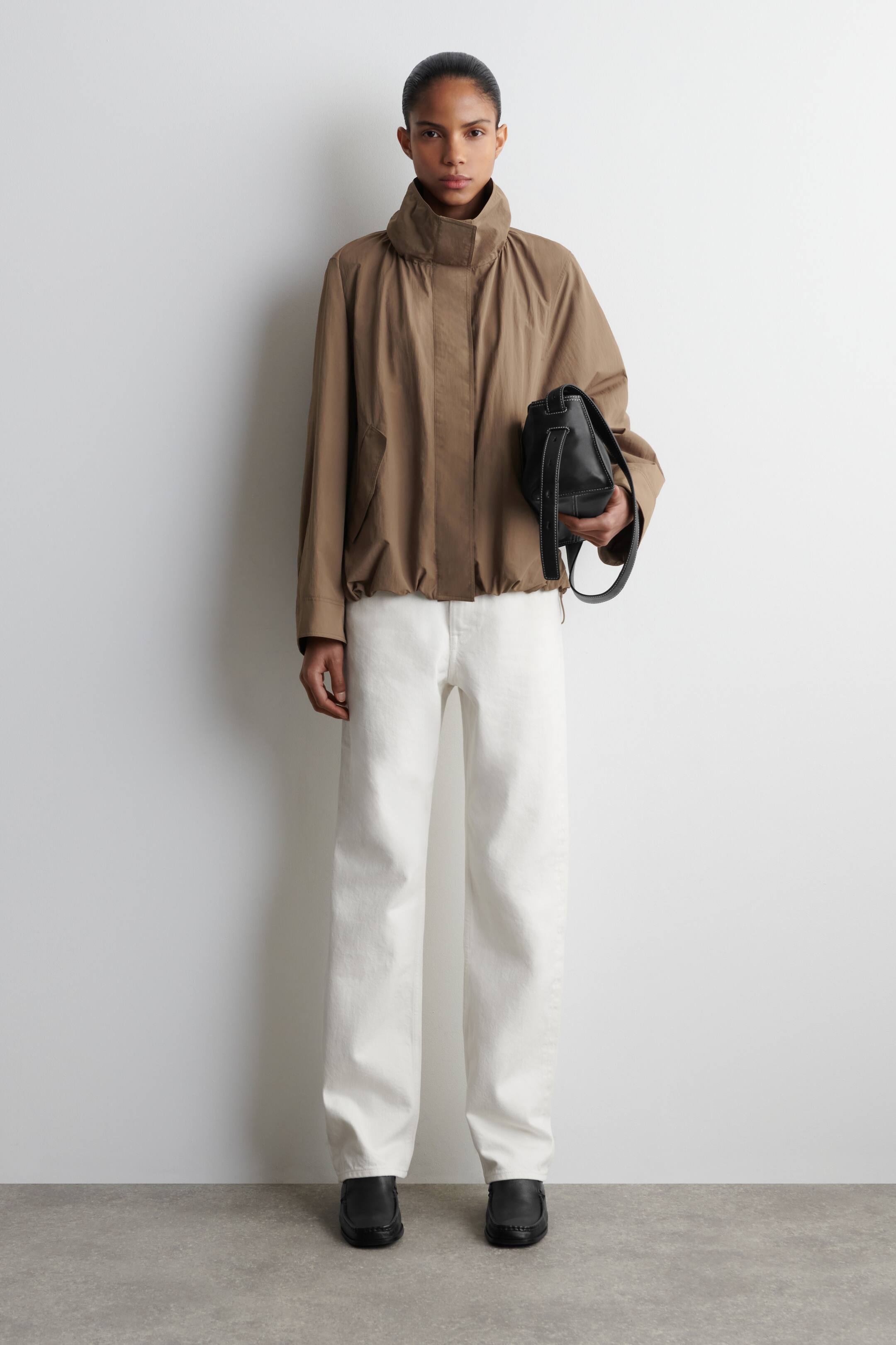 Agrandir l'image: Le produit est présenté en vue de face, mettant en avant une veste ample de couleur brun clair avec un col montant, des manches longues et un ourlet élastiqué. Elle est associée à un pantalon blanc à jambes larges, des chaussures noires à enfiler, et un sac à main structuré noir avec des coutures contrastées blanches.