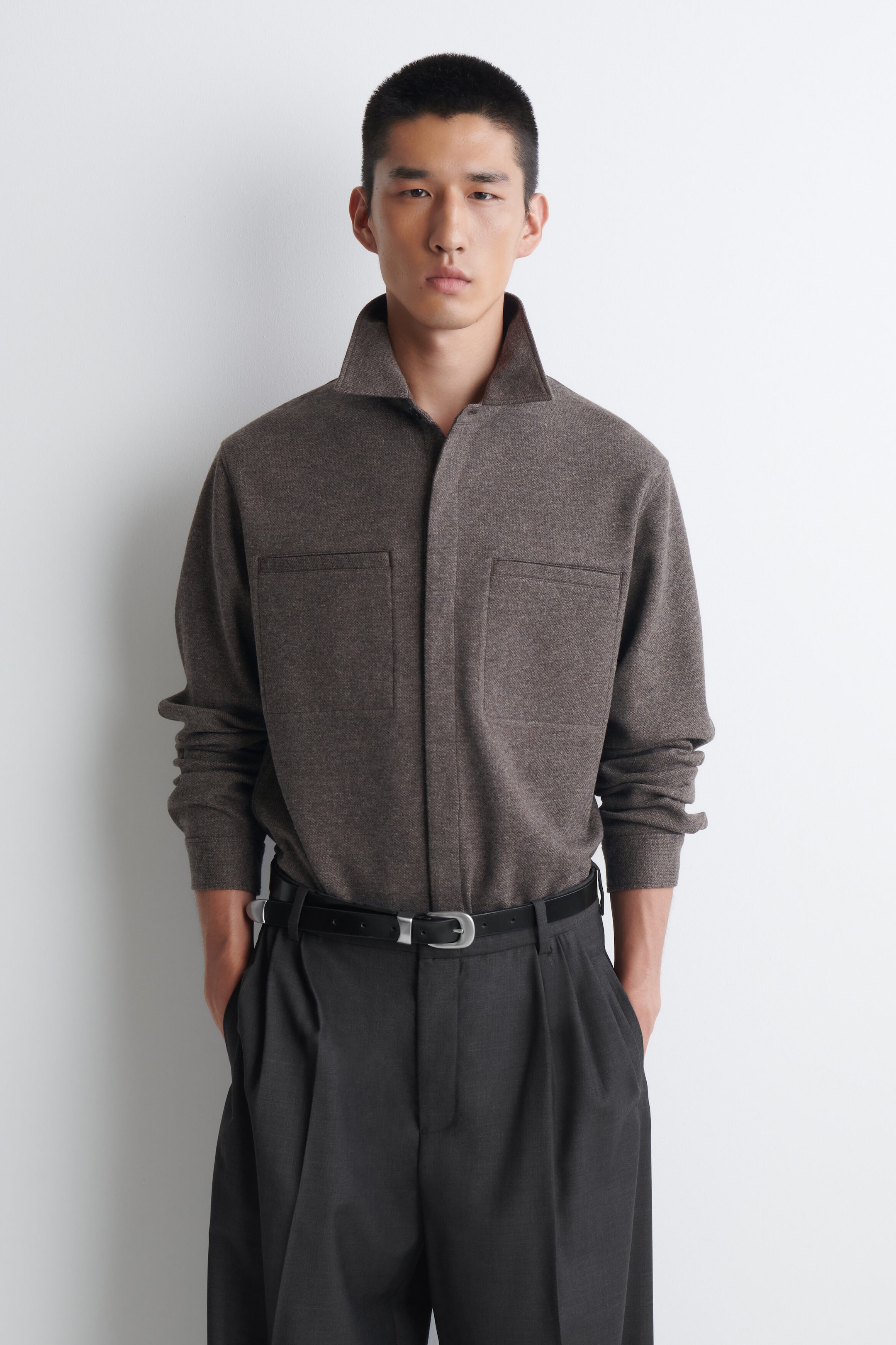 A brown utility overshirt features a textured weave, a spread collar, and two front patch pockets, and is worn tucked into dark trousers. A brown utility overshirt features a textured weave, a spread collar, and two front patch pockets, and is worn tucked into dark trousers.