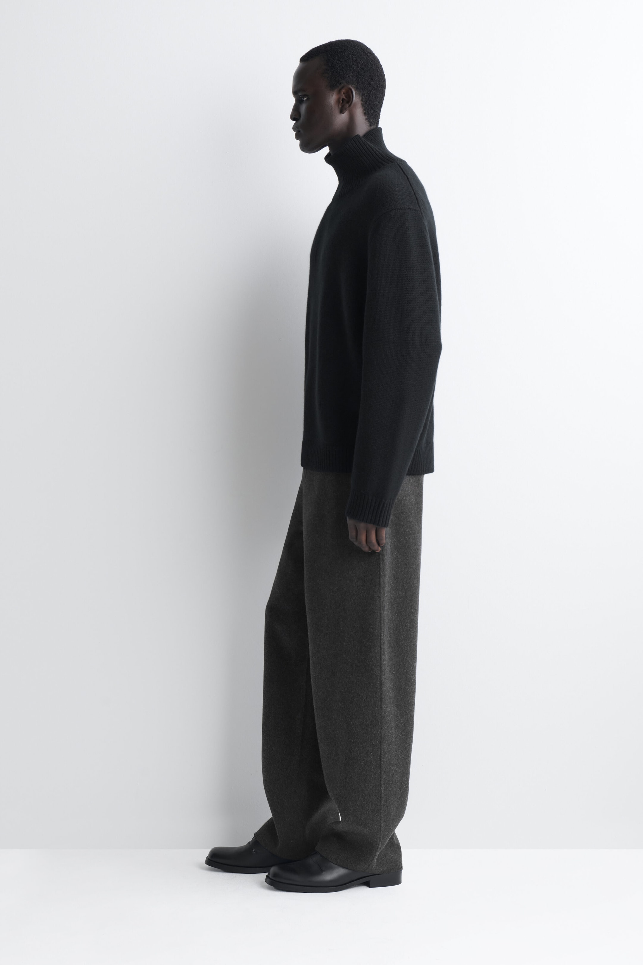 Doppelseitige Wollhose mit Relaxed Fit und weitem Bein in Dunkelgrau, in einer seitlichen Profilansicht nach links gezeigt, wodurch der lockere Fall und die entspannte Silhouette betont werden. Die Hose fällt über schwarze Lederschuhe und wird mit einem schwarzen Strickpullover mit hohem Kragen gestylt. Doppelseitige Wollhose mit Relaxed Fit und weitem Bein in Dunkelgrau, in einer seitlichen Profilansicht nach links gezeigt, wodurch der lockere Fall und die entspannte Silhouette betont werden. Die Hose fällt über schwarze Lederschuhe und wird mit einem schwarzen Strickpullover mit hohem Kragen gestylt.