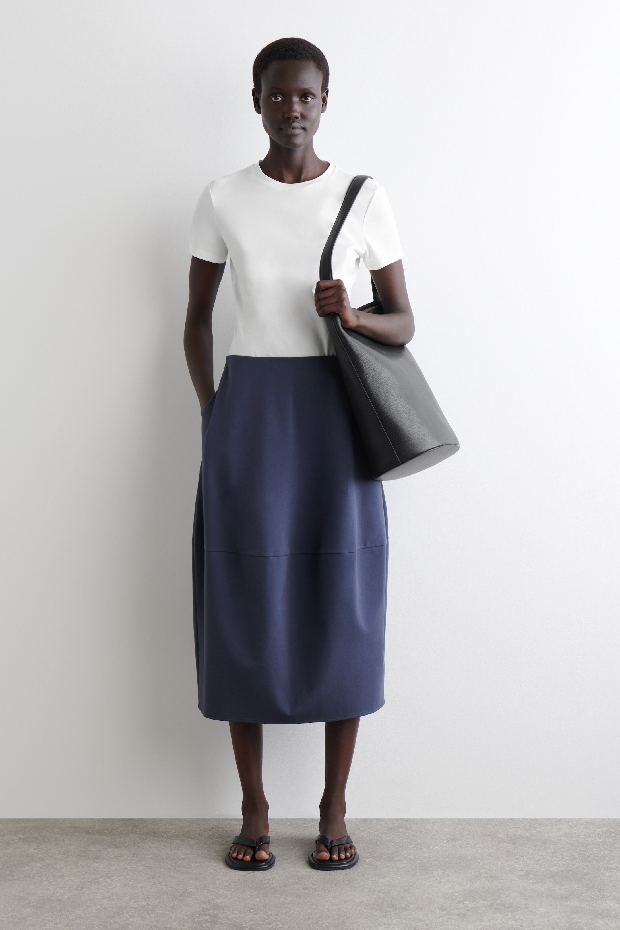 View larger image: A white short-sleeved crew neck t-shirt is being worn, tucked into a navy midi skirt that features side pockets and a horizontal seam detail. A black soft shoulder bag is being carried, and black flip-flops are being worn.