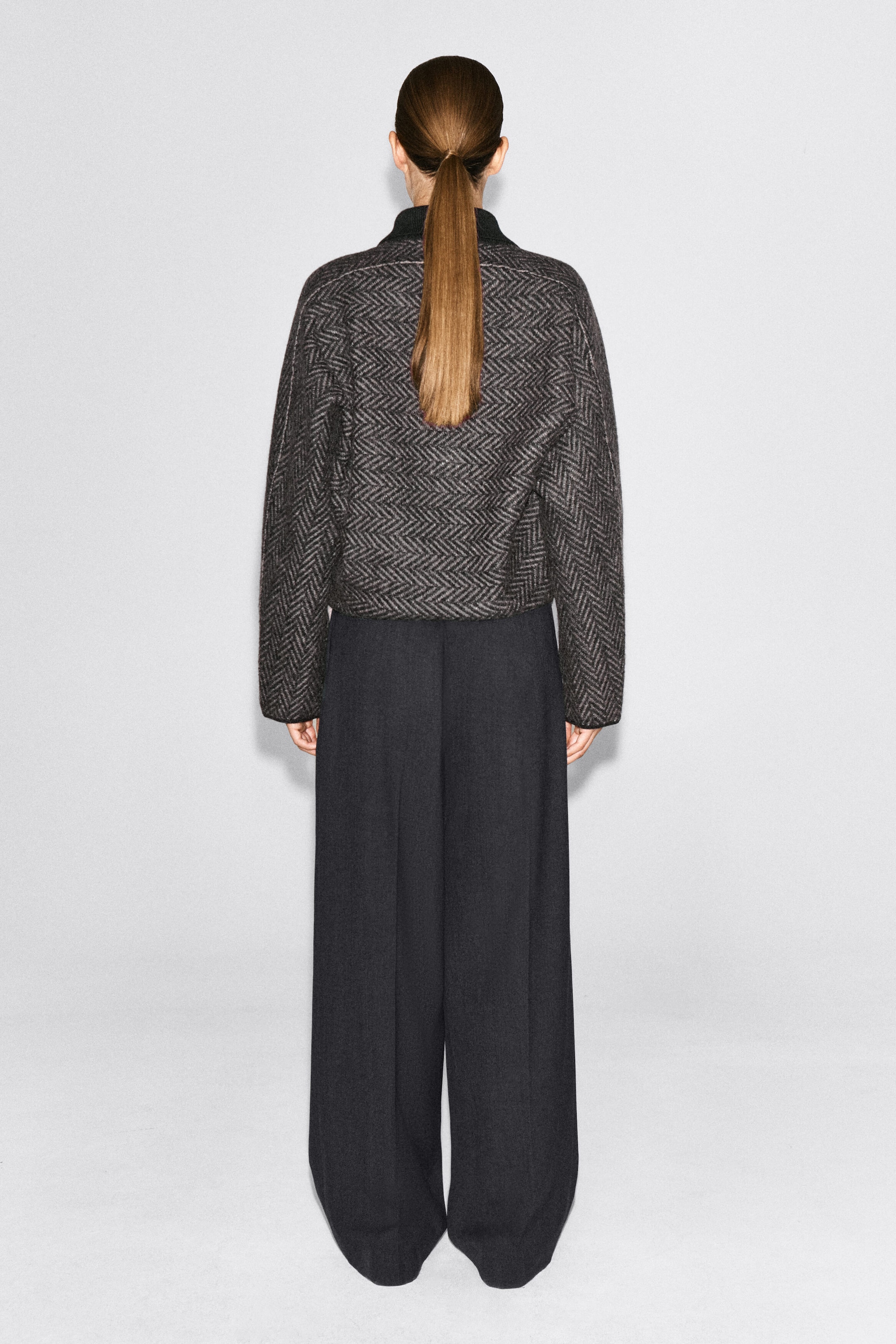 Une veste en tricot à motif chevrons, raccourcie et de couleur gris foncé, est portée par le mannequin, vu de dos. La veste présente une silhouette décontractée, des manches longues et un col côtelé foncé, et elle est agencée à un pantalon foncé à jambe large.