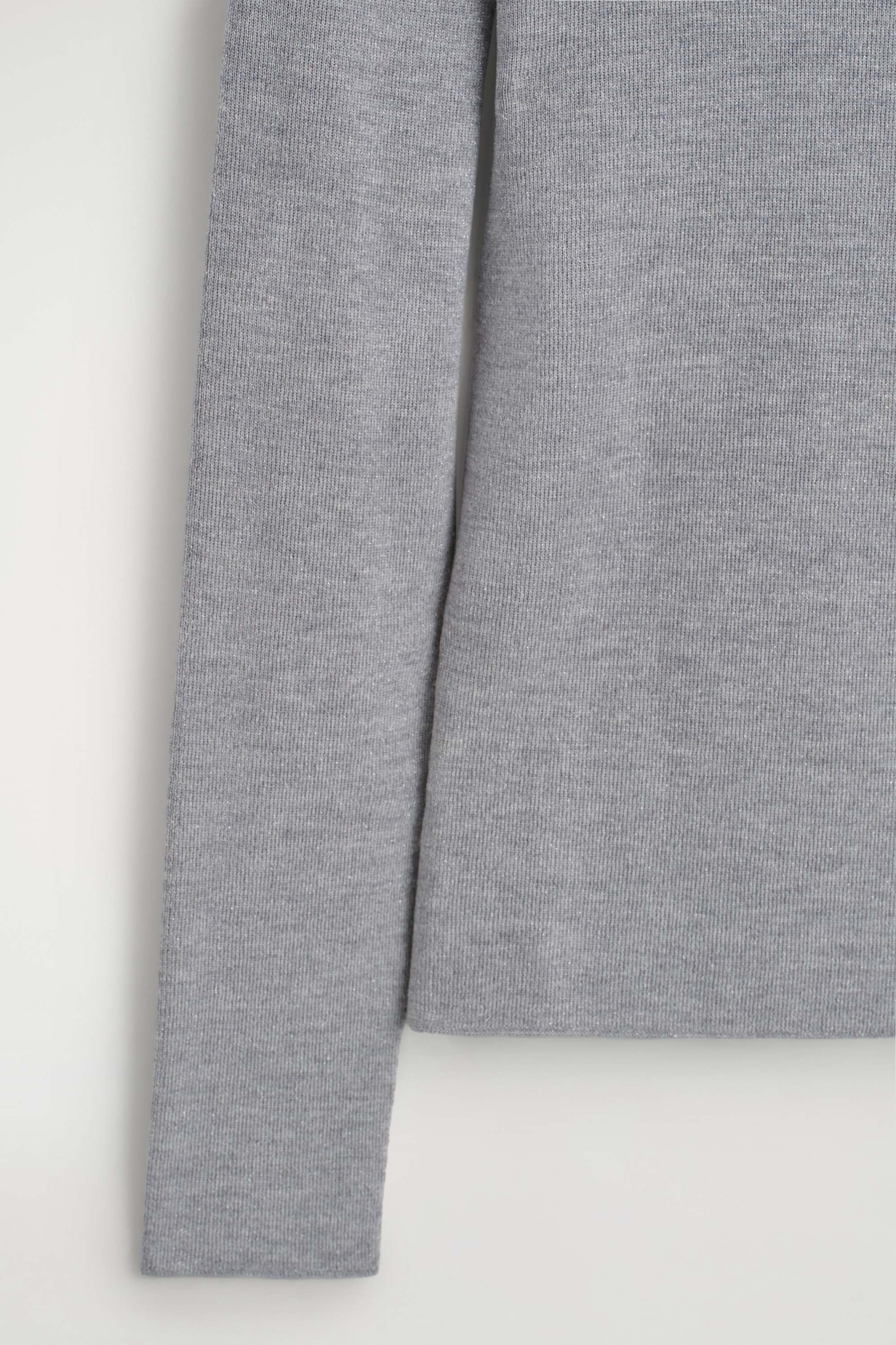 The sparkly seamless wool funnel-neck top showcases its fine-knit, light gray fabric, woven with subtle metallic threads for a shimmering effect. The long sleeve hangs straight down beside the flat, seamless hem of the body.