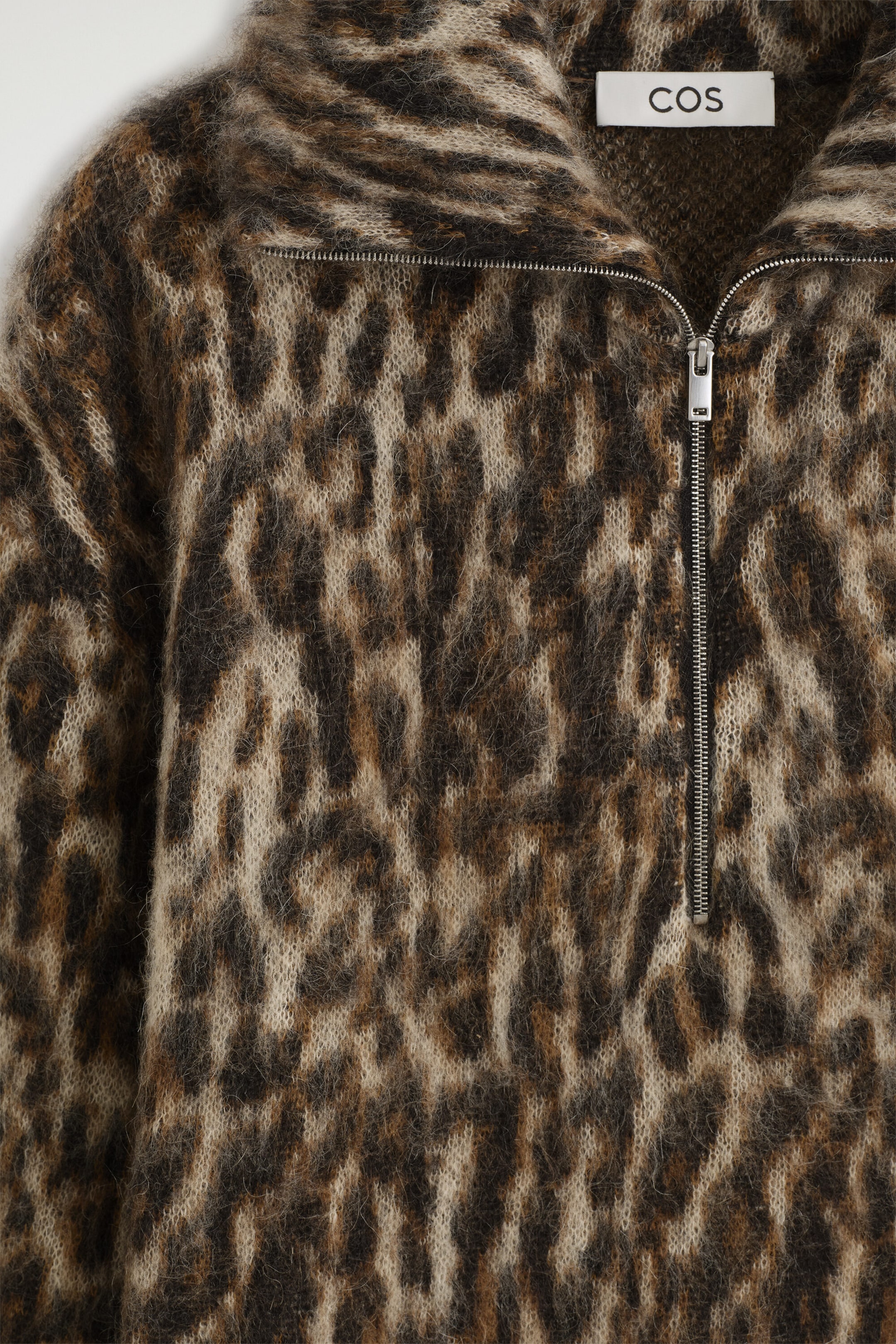 Showing a close-up front view, this fuzzy leopard-print knit jumper features a brushed texture in shades of brown, black and beige. The jumper displays a silver half-zip fastening at the funnel neck, revealing a white brand label sewn inside. Showing a close-up front view, this fuzzy leopard-print knit jumper features a brushed texture in shades of brown, black and beige. The jumper displays a silver half-zip fastening at the funnel neck, revealing a white brand label sewn inside.