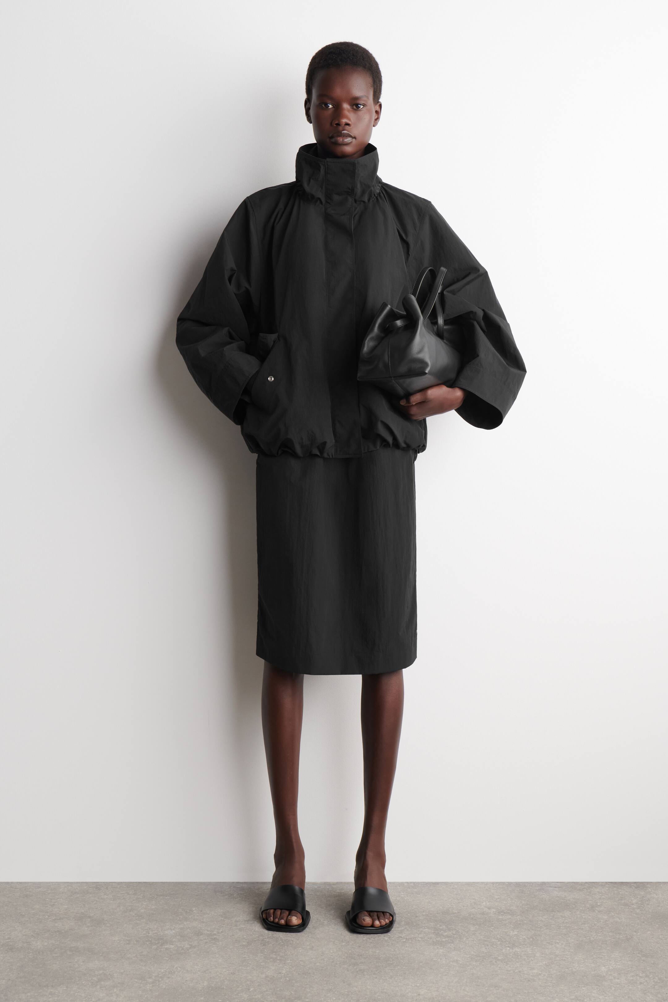 View larger image: A black jacket featuring a high stand collar, dropped shoulders, voluminous sleeves with snap-button cuffs, and an elasticated hem. It is styled with a midi-length black straight-fit skirt, a black soft-structured handbag, and black flat slide sandals.