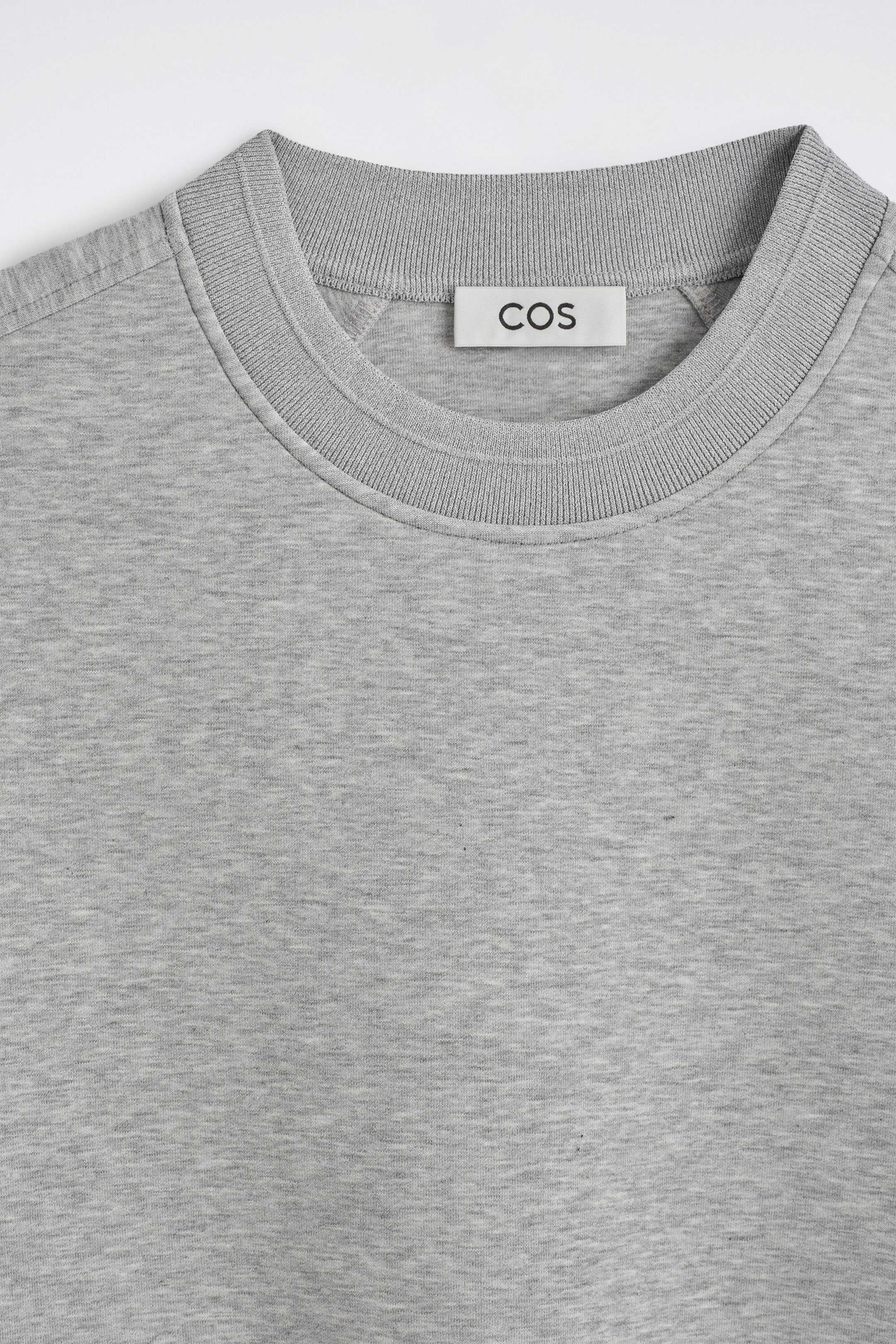 The relaxed scuba sweatshirt is displaying a close-up front view of its textured heather gray mélange fabric and thick ribbed crew neck. A white rectangular label featuring the brand name "COS" is showing inside the collar line near the raglan shoulder seams. The relaxed scuba sweatshirt is displaying a close-up front view of its textured heather gray mélange fabric and thick ribbed crew neck. A white rectangular label featuring the brand name "COS" is showing inside the collar line near the raglan shoulder seams.
