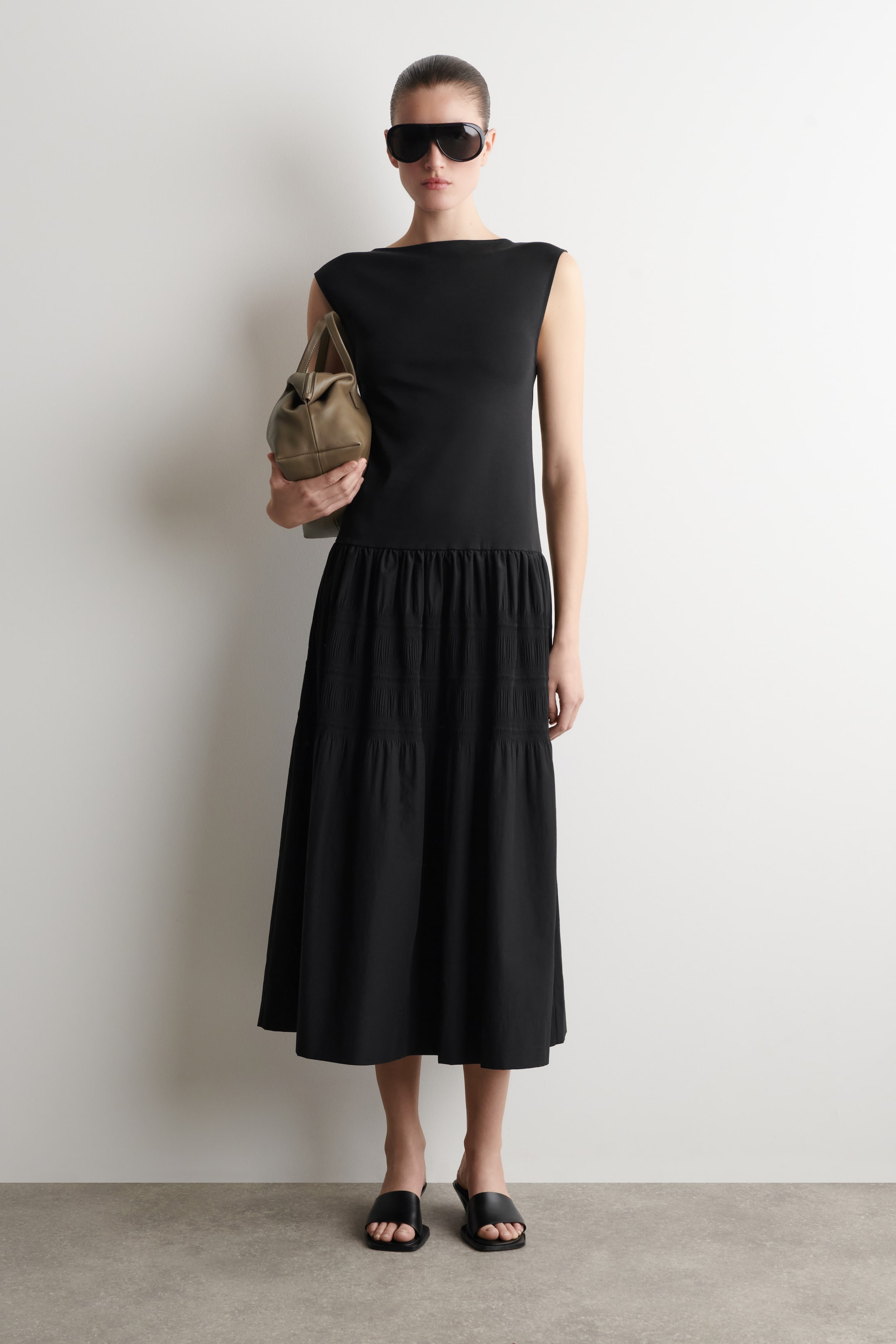 Grösseres Bild ansehen: Ein schwarzes Midikleid, das ein ärmelloses Jersey-Oberteil und einen gewebten Rock mit taktilem Zickzack-Rauchmuster auf der oberen Stufe aufweist, wird vom Model getragen. Das Model hält zudem eine gedämpft grüne Handtasche und trägt schwarze Slides sowie eine Sonnenbrille.