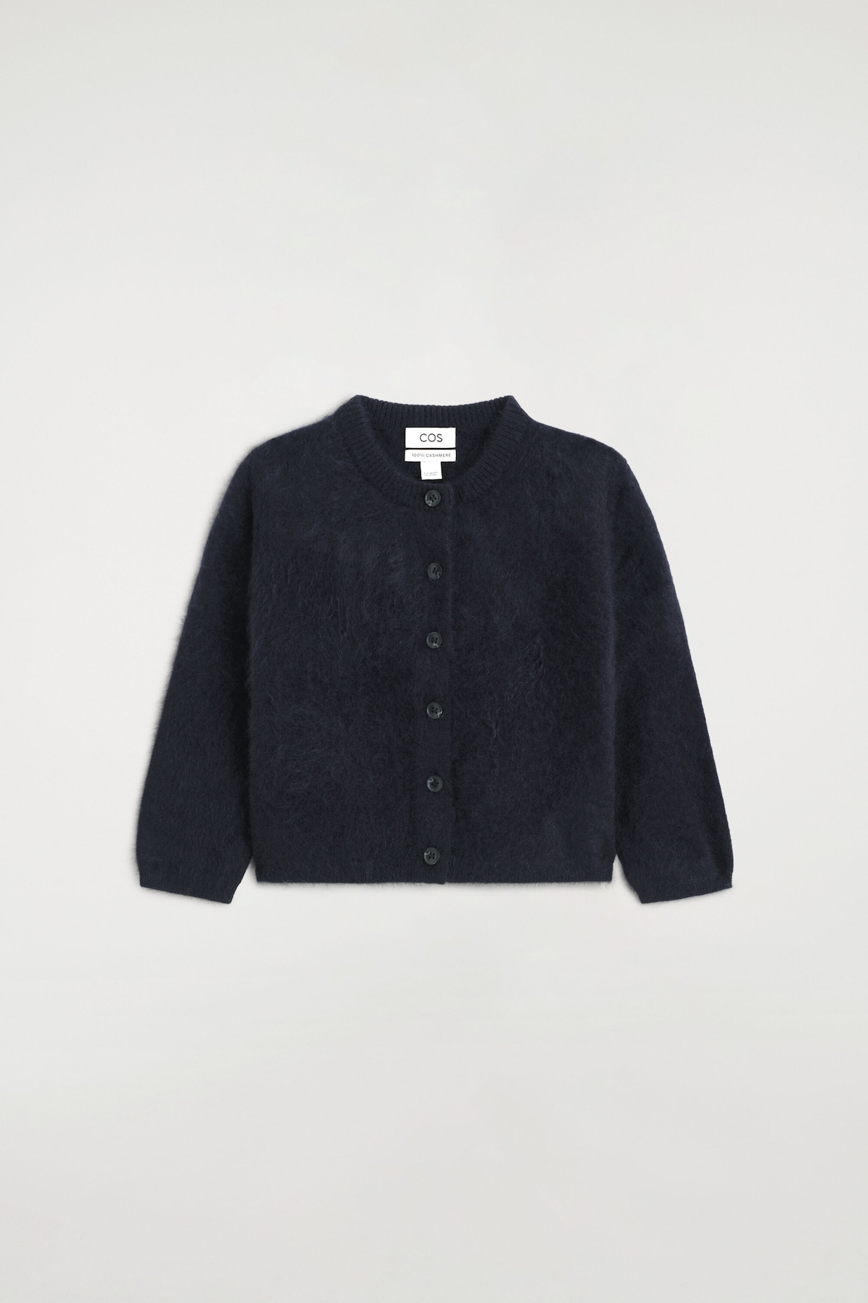 The kids’ brushed pure cashmere cardigan is shown from the front, featuring a dark navy blue color and a soft, fuzzy brushed texture. It is designed with a ribbed crew neck, a central button-down closure with six dark buttons, and long sleeves.