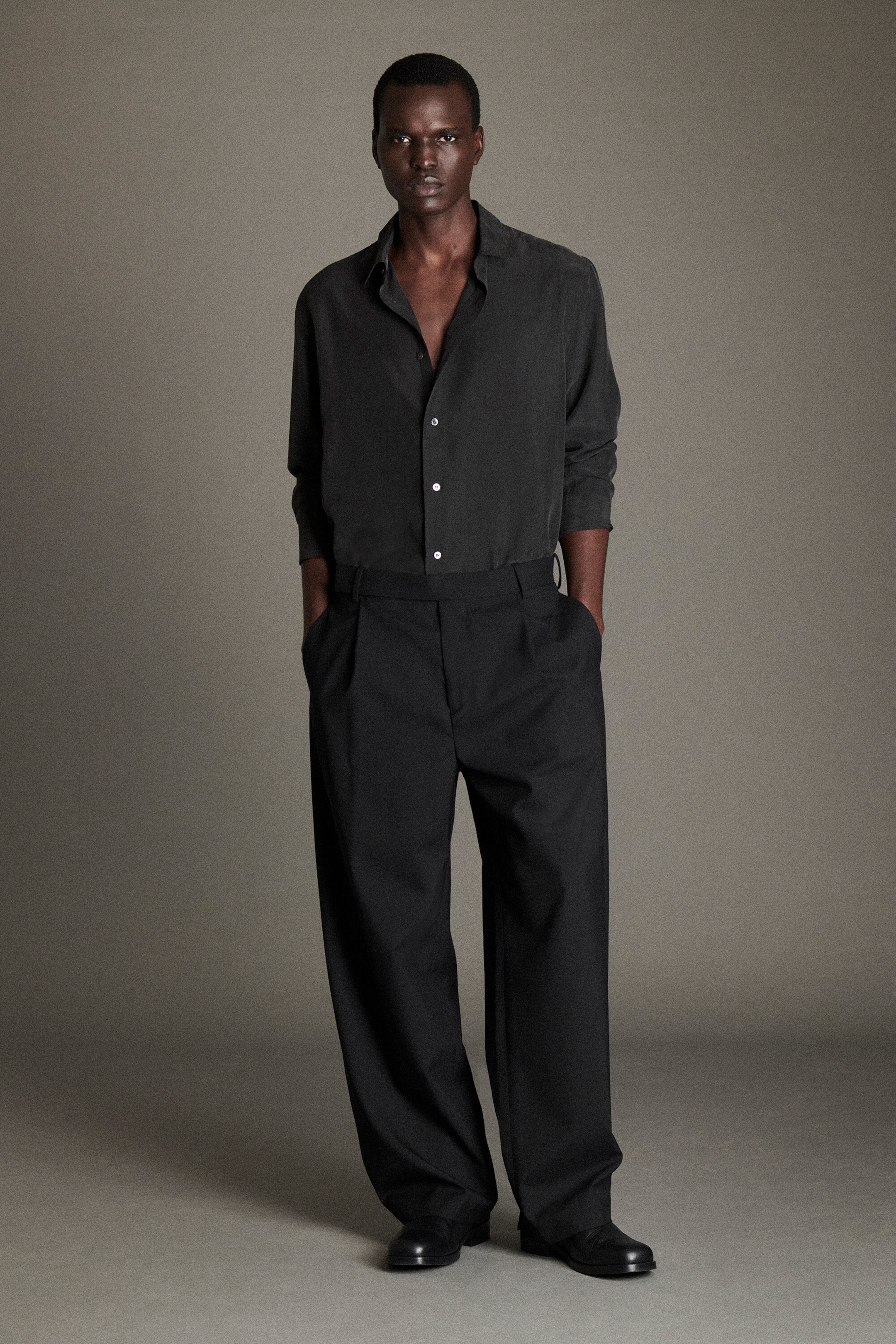 A model is standing facing the front, wearing a charcoal-grey sandwashed silk shirt with a relaxed, partially unbuttoned collar and rolled-up sleeves. The shirt is tucked into a pair of black wool relaxed-fit barrel-leg trousers featuring front pleats, side slip pockets, and a wide, slightly curved silhouette. A model is standing facing the front, wearing a charcoal-grey sandwashed silk shirt with a relaxed, partially unbuttoned collar and rolled-up sleeves. The shirt is tucked into a pair of black wool relaxed-fit barrel-leg trousers featuring front pleats, side slip pockets, and a wide, slightly curved silhouette.