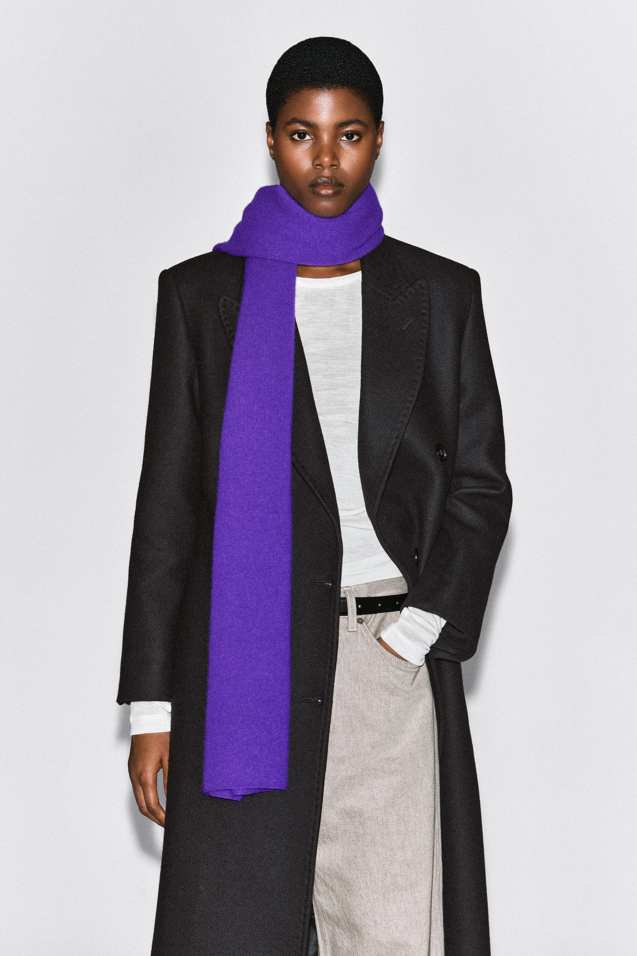 A purple cashmere scarf is worn by the model, wrapped once around the neck with one long end hanging down the front. The scarf is layered over a black double-breasted coat, a white top, and light gray trousers. A purple cashmere scarf is worn by the model, wrapped once around the neck with one long end hanging down the front. The scarf is layered over a black double-breasted coat, a white top, and light gray trousers.