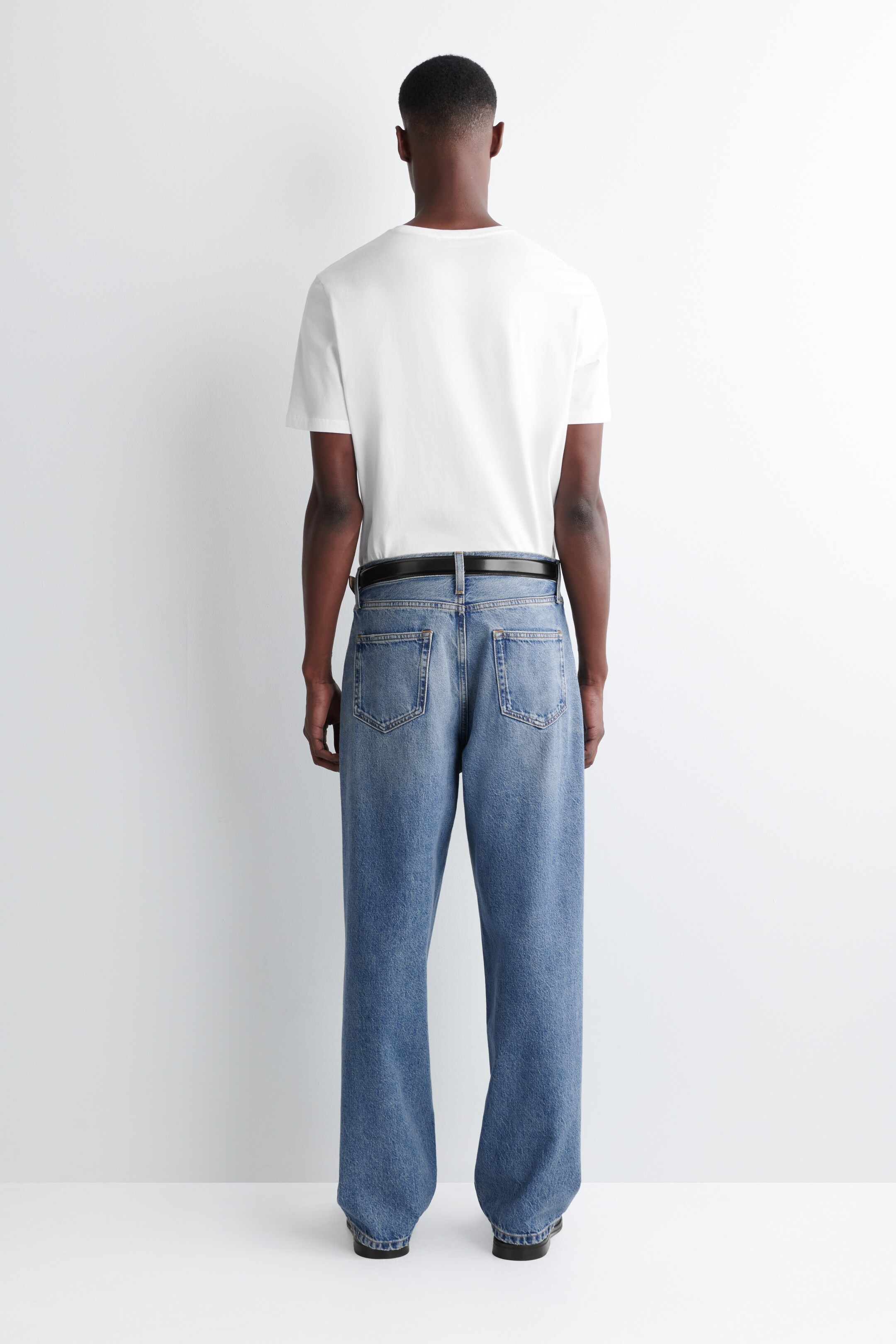 Visa större bild: En enkel vit t-shirt med korta ärmar och ljusblå tvättade jeans bärs av en person sedd bakifrån. Jeansen har en avslappnad, rak passform med synliga bakfickor och bälteshällor, genom vilka ett svart bälte är trätt.