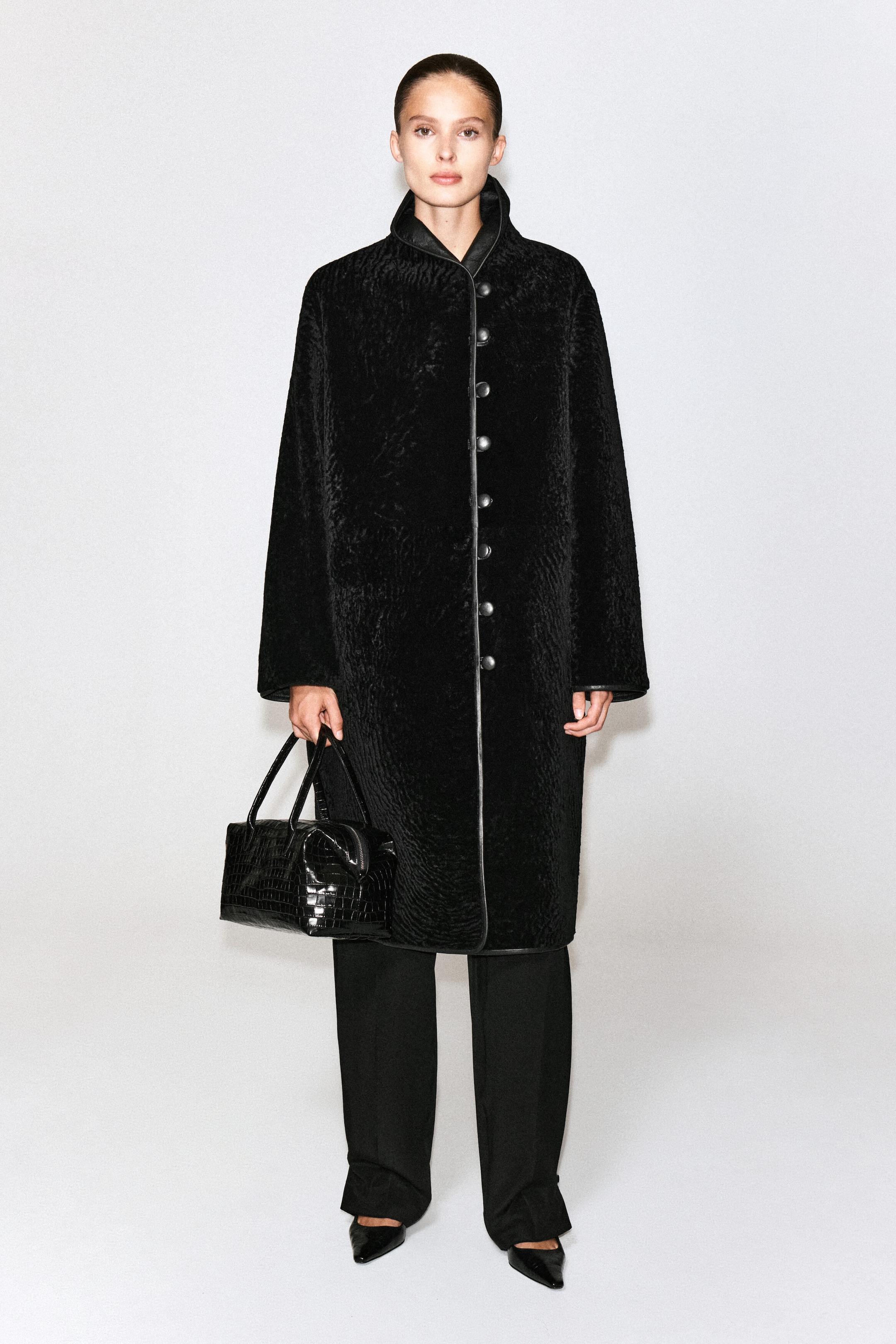A black, croc-debossed leather bowling bag with a structured, sculptural shape and double top handles is being held by the model. The model is carrying the bag in one hand while wearing a long, textured black coat and black trousers. A black, croc-debossed leather bowling bag with a structured, sculptural shape and double top handles is being held by the model. The model is carrying the bag in one hand while wearing a long, textured black coat and black trousers.