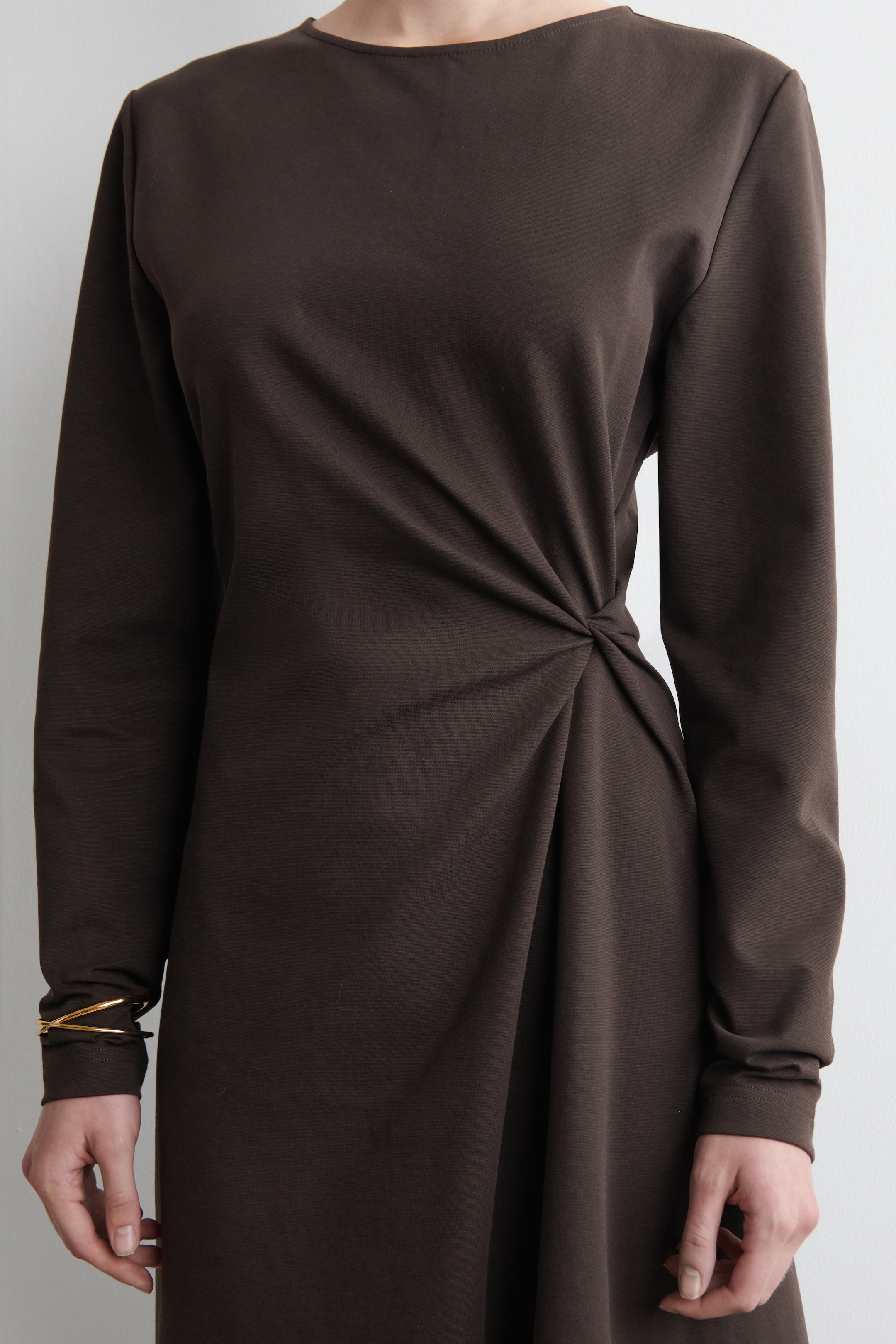 Agrandir l'image: Une robe à manches longues de couleur brun foncé présente un col rond. Le tissu se rassemble du côté gauche de la taille avec un détail noué, créant des drapés et des plis diagonaux sur le devant de la robe.