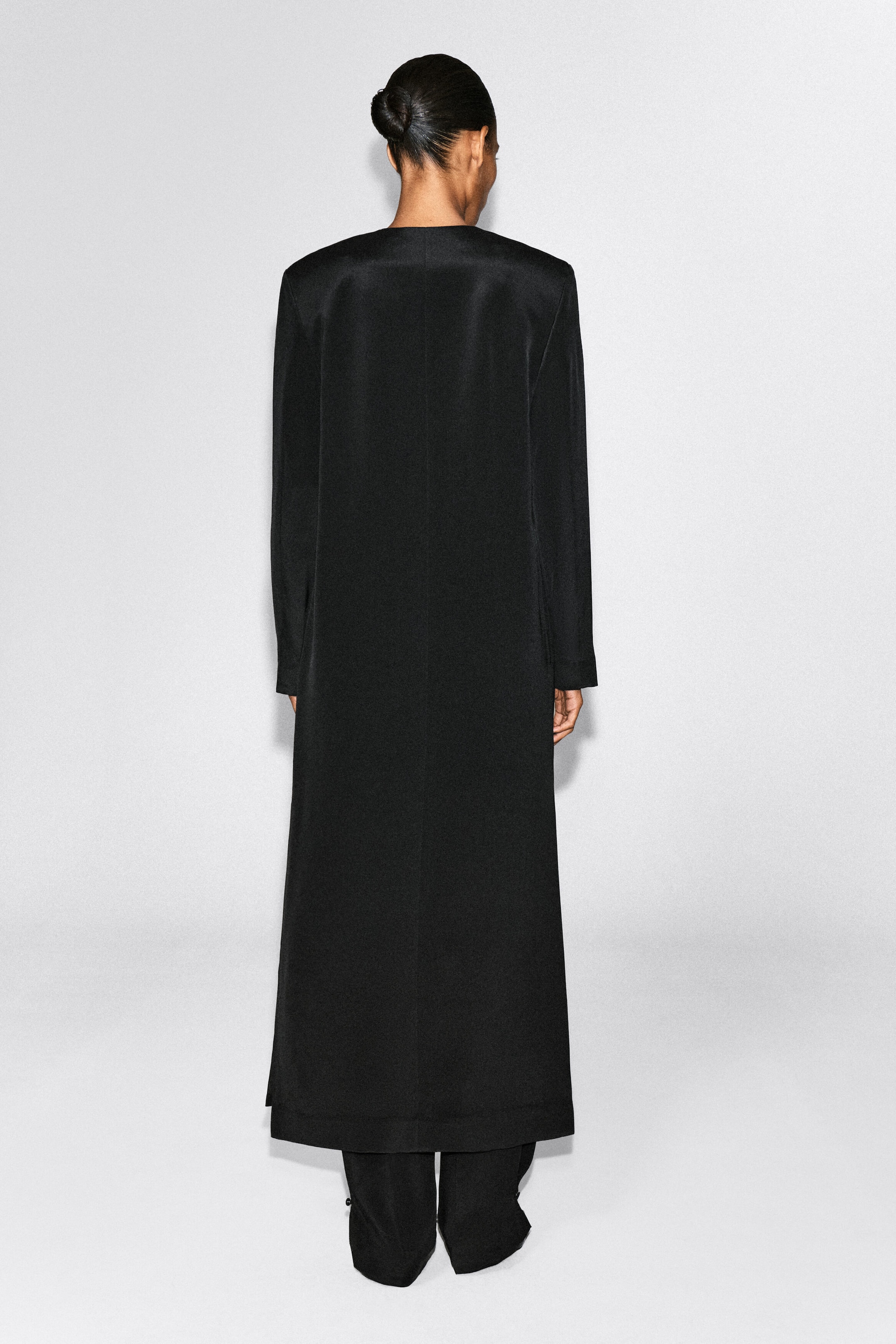 Das Model trägt einen schwarzen, kragenlosen Maxi-Mantel, von hinten gezeigt, um seine lange, gerade Silhouette und die glatte Textur hervorzuheben. Der Mantel ist mit einer passenden schwarzen Hose kombiniert. Das Model trägt einen schwarzen, kragenlosen Maxi-Mantel, von hinten gezeigt, um seine lange, gerade Silhouette und die glatte Textur hervorzuheben. Der Mantel ist mit einer passenden schwarzen Hose kombiniert.