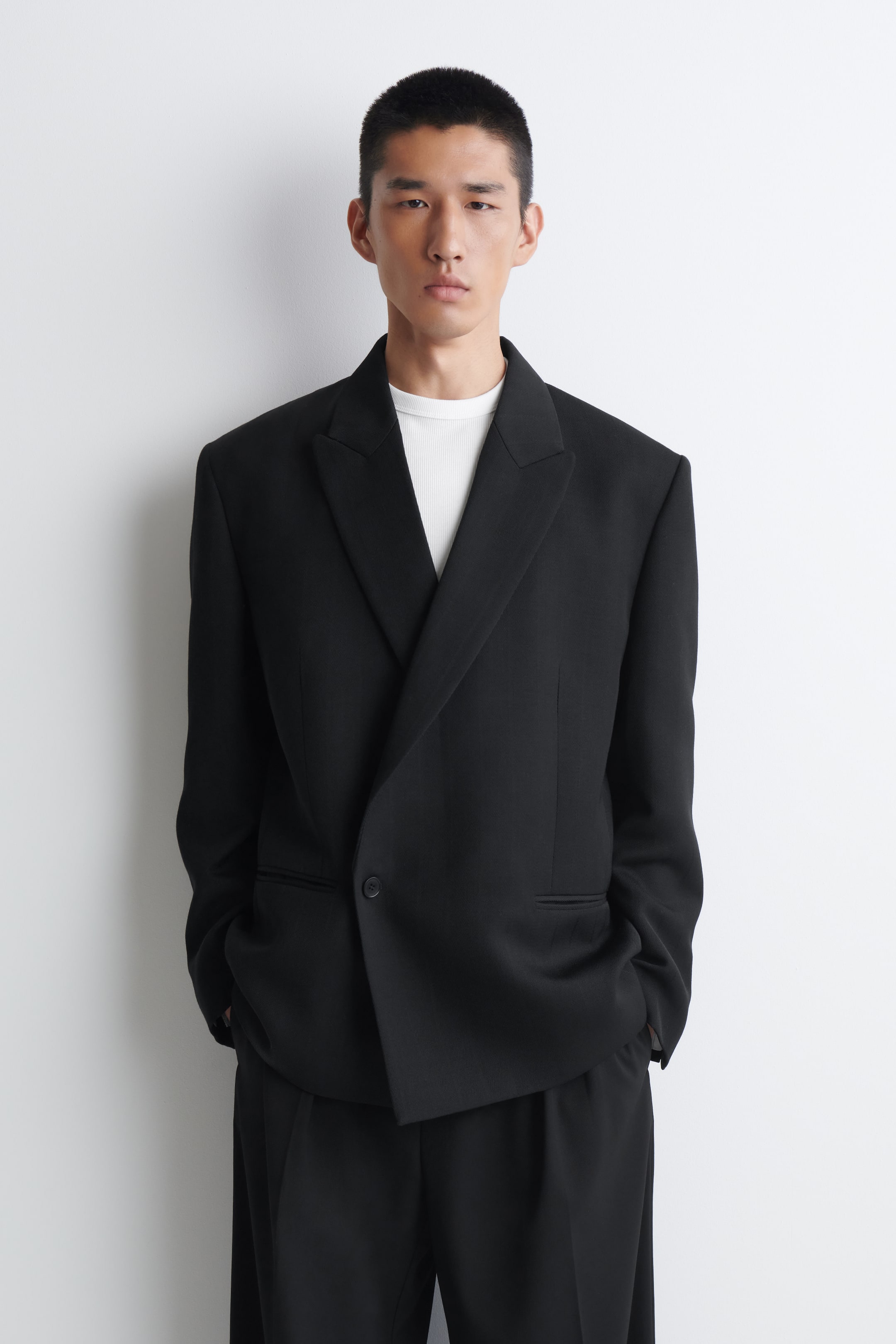 Größeres Bild anzeigen: Der schwarze Zweireiher-Blazer aus Twill präsentiert sich von vorne in einer entspannten Silhouette mit strukturierten Schultern, breiten Peak-Lapels und einem Ein-Knopf-Verschluss. Dazu wird ein weißes T-Shirt mit Rundhalsausschnitt getragen, kombiniert mit passender schwarzer Hose.