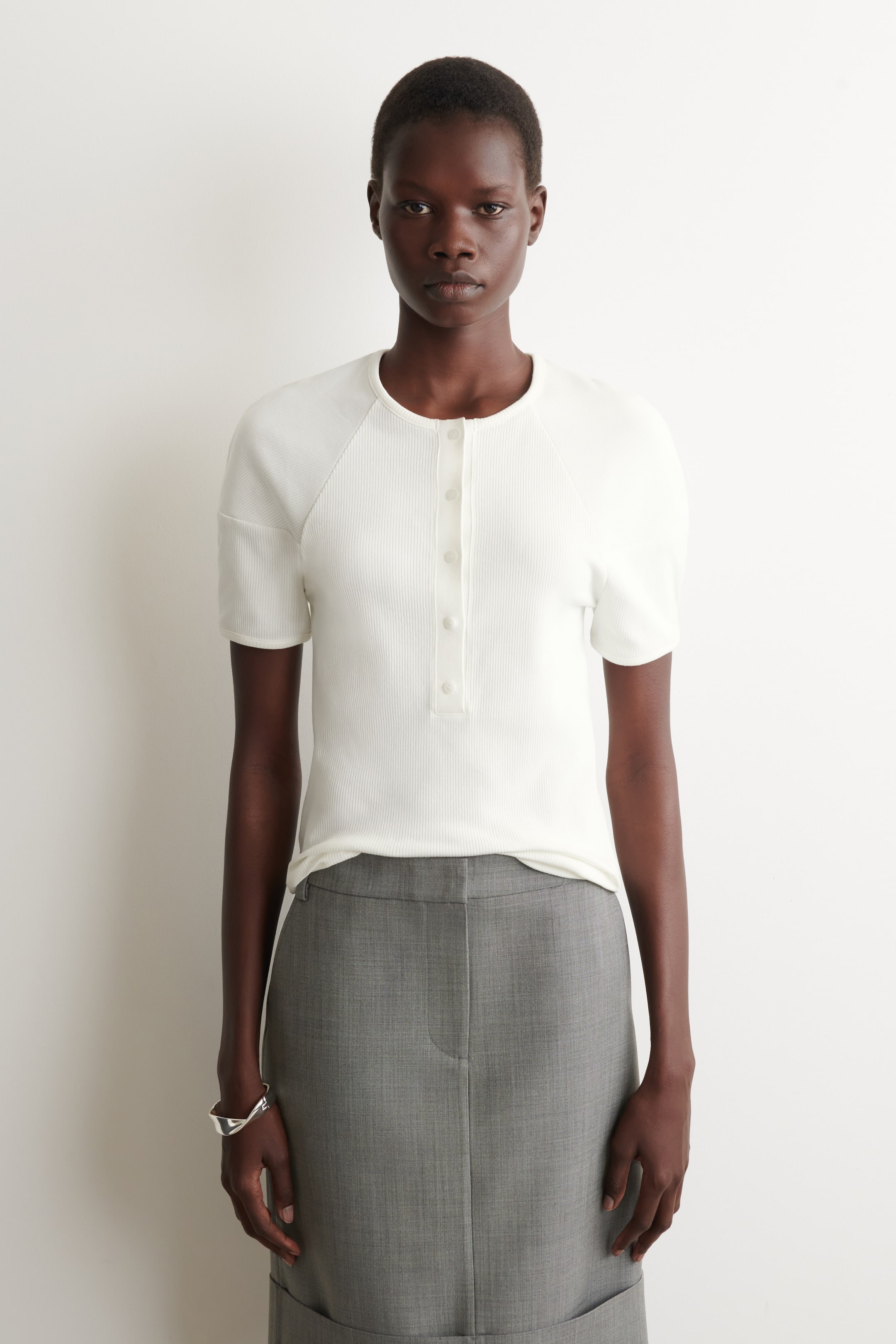Zobraziť väčší obrázok: Biele rebrované tričko s krátkymi rukávmi je otočené dopredu, s okrúhlym výstrihom a päťgombíkovou Henley légou. Je navrhnuté s raglánovými rukávmi a predstavuje priliehavú siluetu zastrčenú do sivé sukne.
