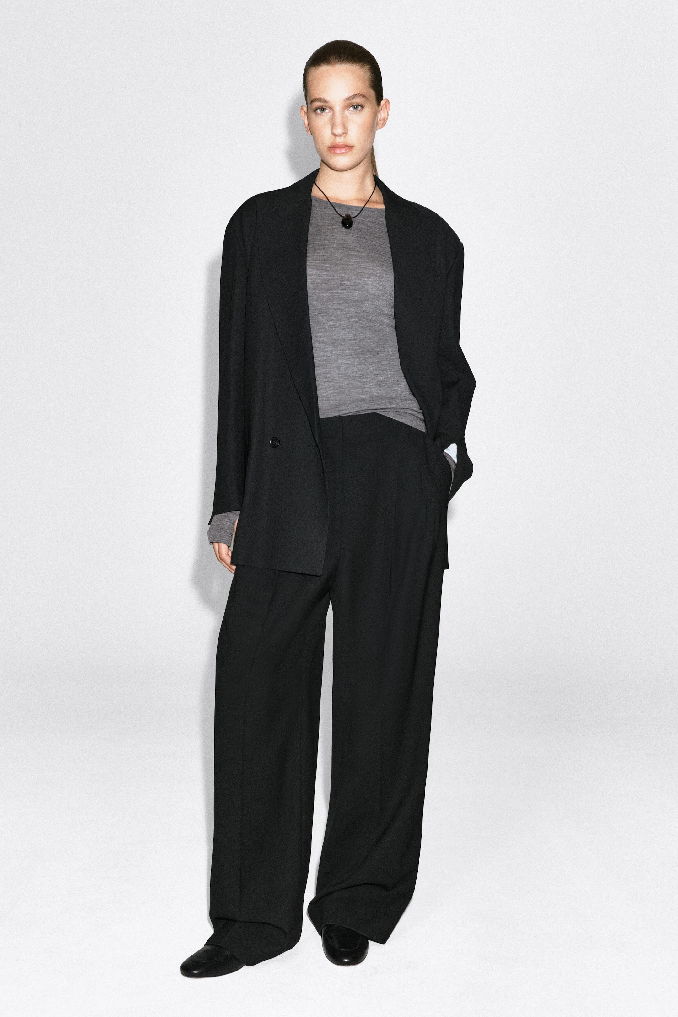 A slim-fit, fine-knit grey merino wool top with a round neckline is worn by the model, layered under an open black blazer and tucked into black wide-leg trousers. The long sleeves of the top extend slightly past the blazer cuffs.