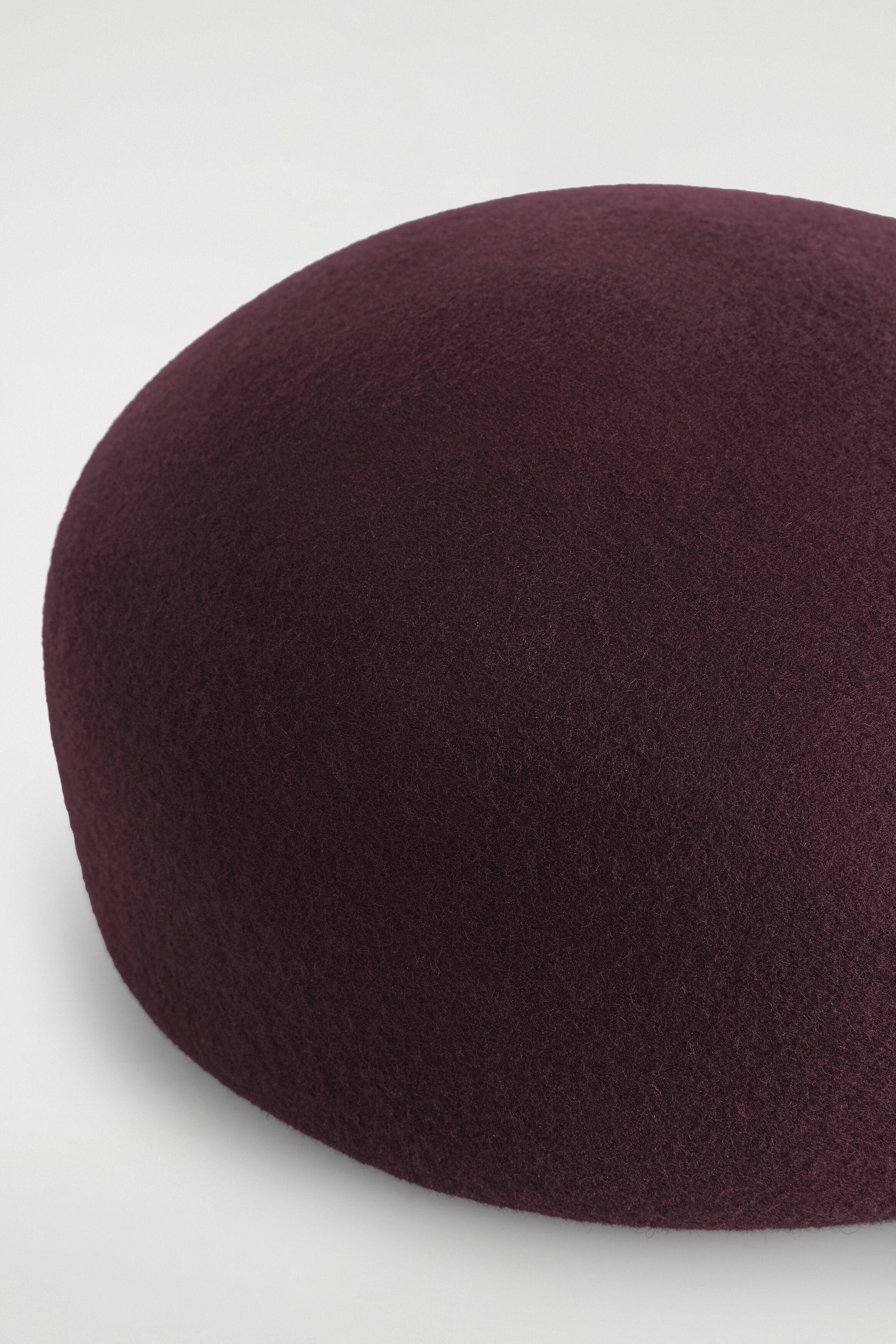 A structured beret in a deep burgundy hue is showcasing its rounded, sculptural shape and soft, textured boiled wool material in a close-up view. A structured beret in a deep burgundy hue is showcasing its rounded, sculptural shape and soft, textured boiled wool material in a close-up view.