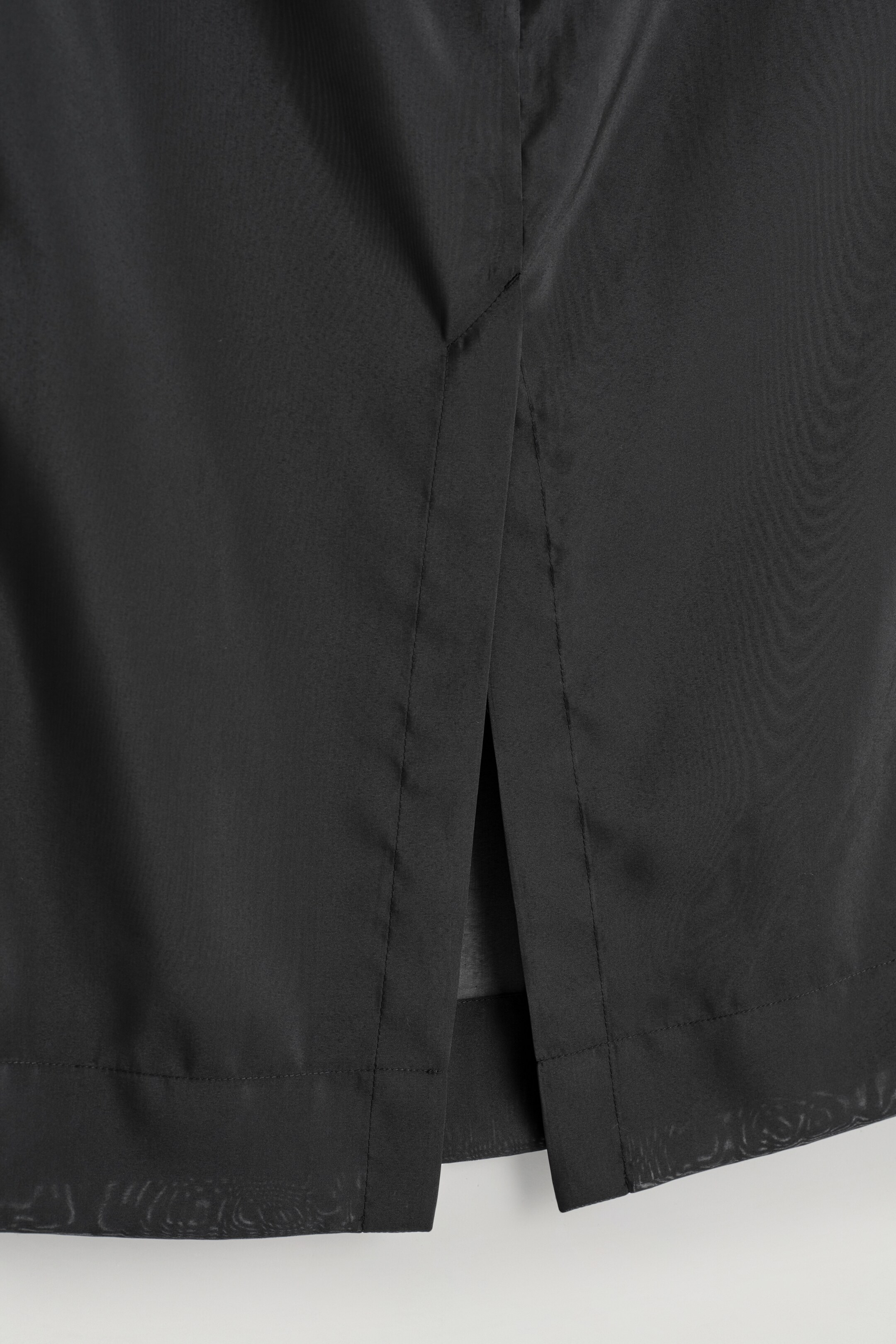 View larger image: A black column skirt is presented from the back, featuring a central vent and a sheer outer layer of gauzy fabric over a less transparent internal lining, visible at the hem.