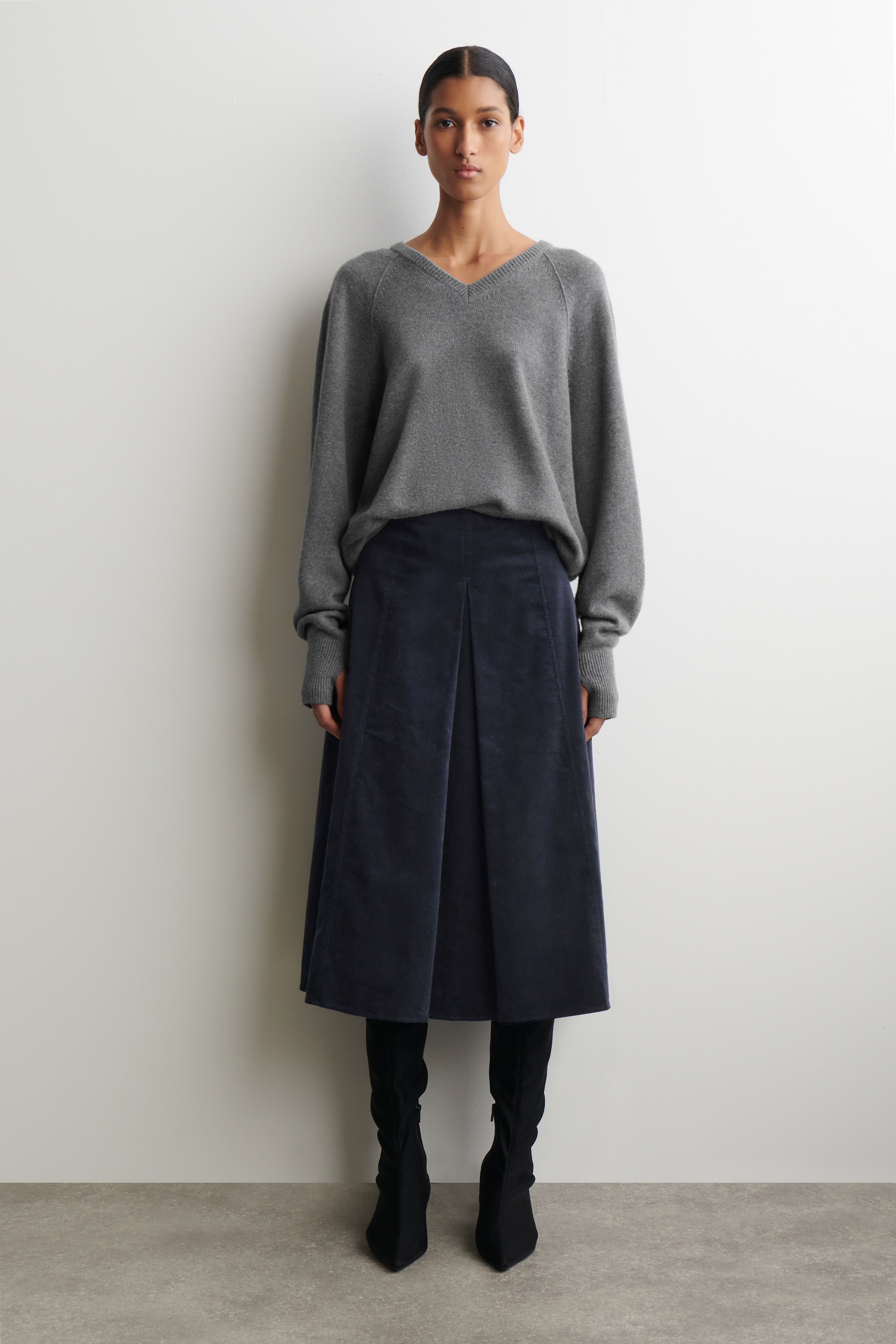 Agrandir l'image: Un pull gris à col en V avec des manches raglan et des poignets côtelés est porté, apparaissant de face et reposant sur une jupe midi en velours côtelé bleu marine. La jupe présente des plis creux à l'avant, et des bottes noires à bout pointu sont également portées en dessous.