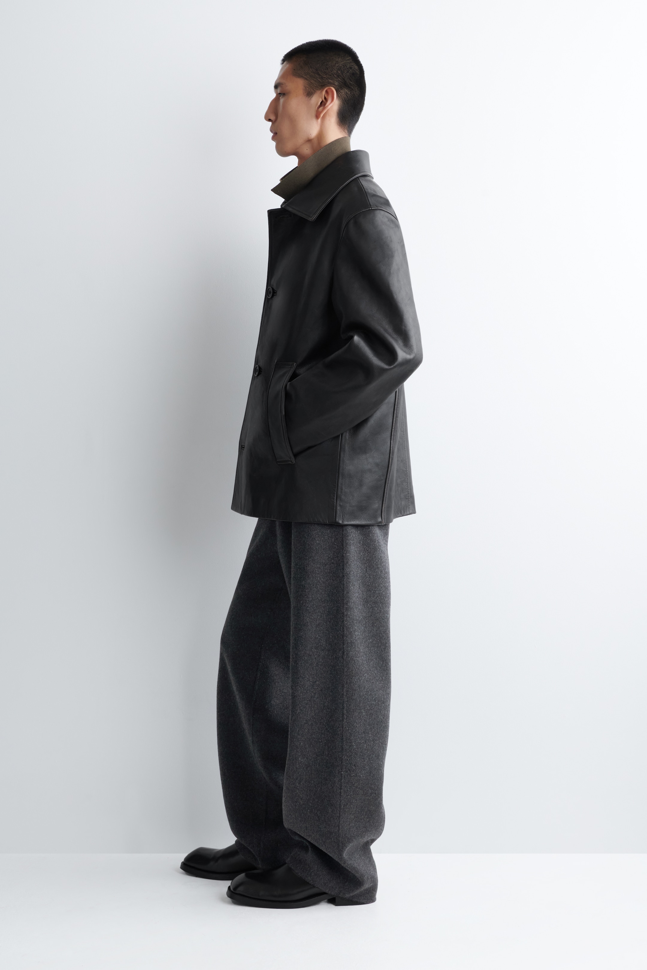 Agrandir l'image: Une veste en similicuir noir, avec un col classique et une fermeture à boutons, est présentée de profil. Elle est superposée à un haut à col montant vert olive et associée à un pantalon large gris foncé qui s'accumule sur des chaussures noires.