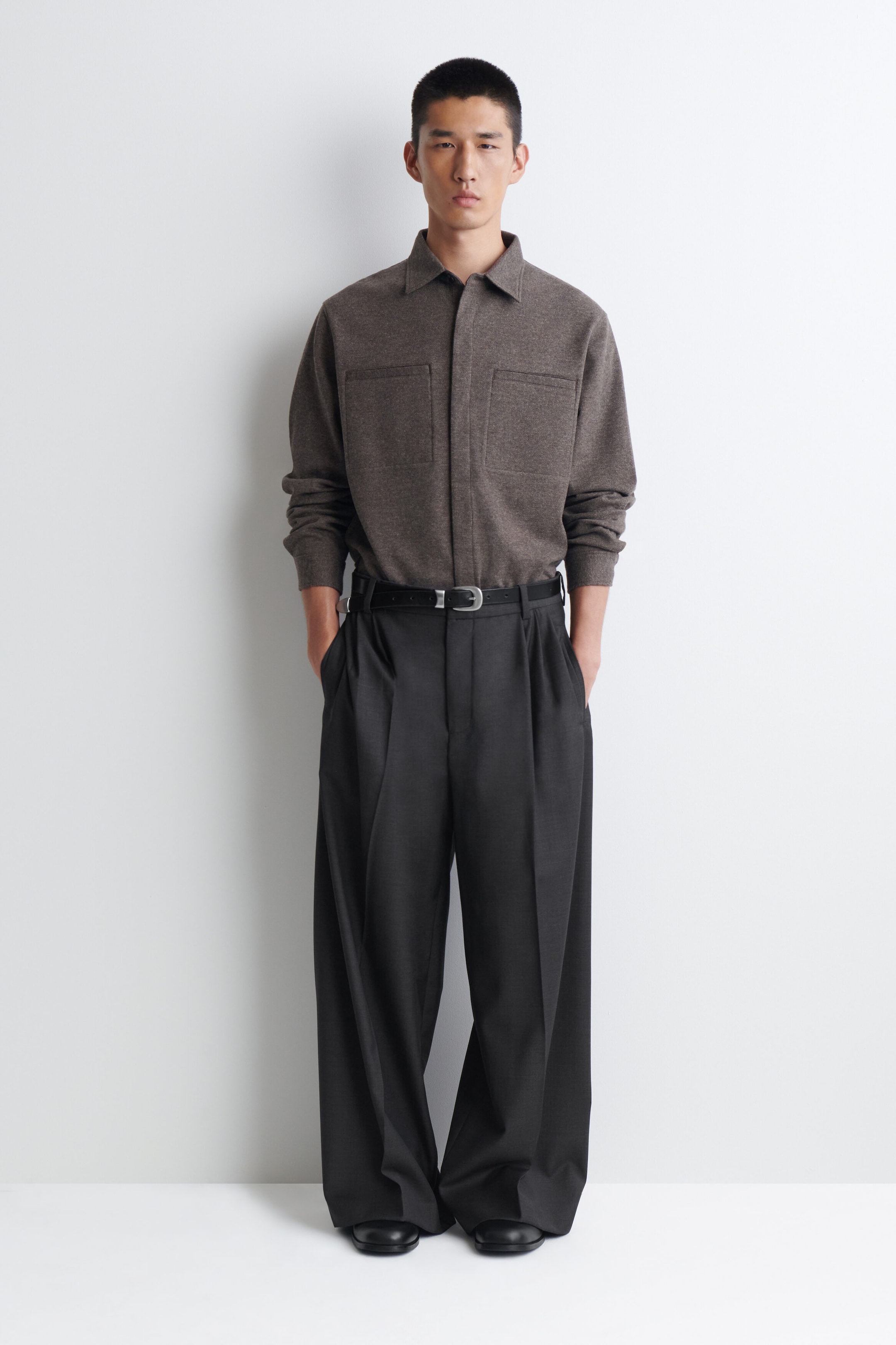 This outfit features a brown knit shirt with a collar, long sleeves, and two chest patch pockets. It is styled with dark grey wide-leg pleated trousers, secured by a black belt with a silver-tone buckle, and black square-toe shoes. This outfit features a brown knit shirt with a collar, long sleeves, and two chest patch pockets. It is styled with dark grey wide-leg pleated trousers, secured by a black belt with a silver-tone buckle, and black square-toe shoes.