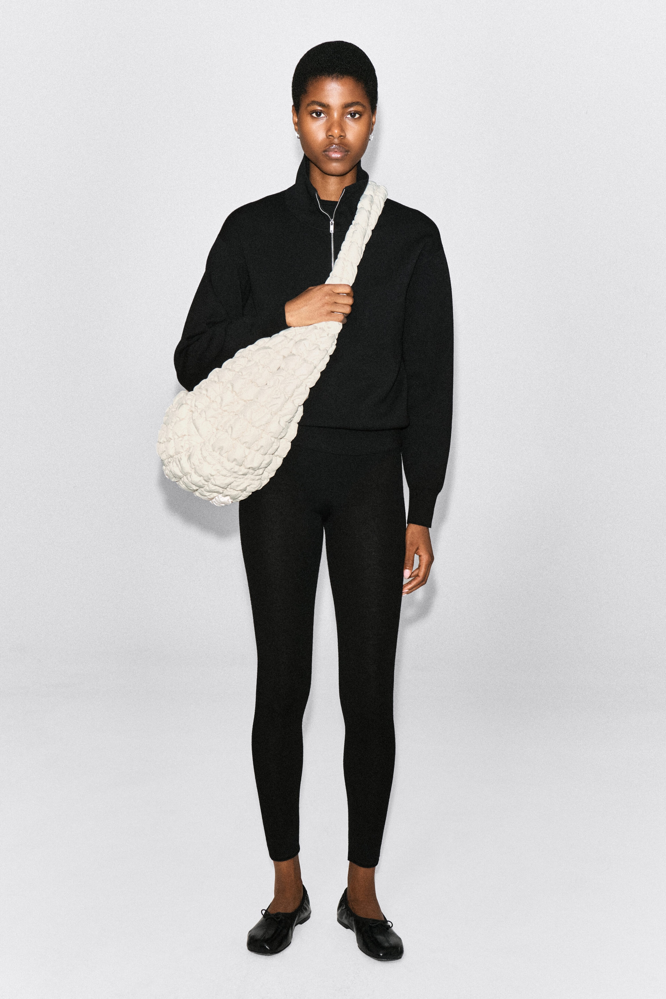 An oversized, off-white quilted crossbody bag featuring a highly textured, puckered finish is worn across the chest by the model. The model is holding the bag's wide strap with one hand, styled over a black zip-up sweater and matching leggings. An oversized, off-white quilted crossbody bag featuring a highly textured, puckered finish is worn across the chest by the model. The model is holding the bag's wide strap with one hand, styled over a black zip-up sweater and matching leggings.