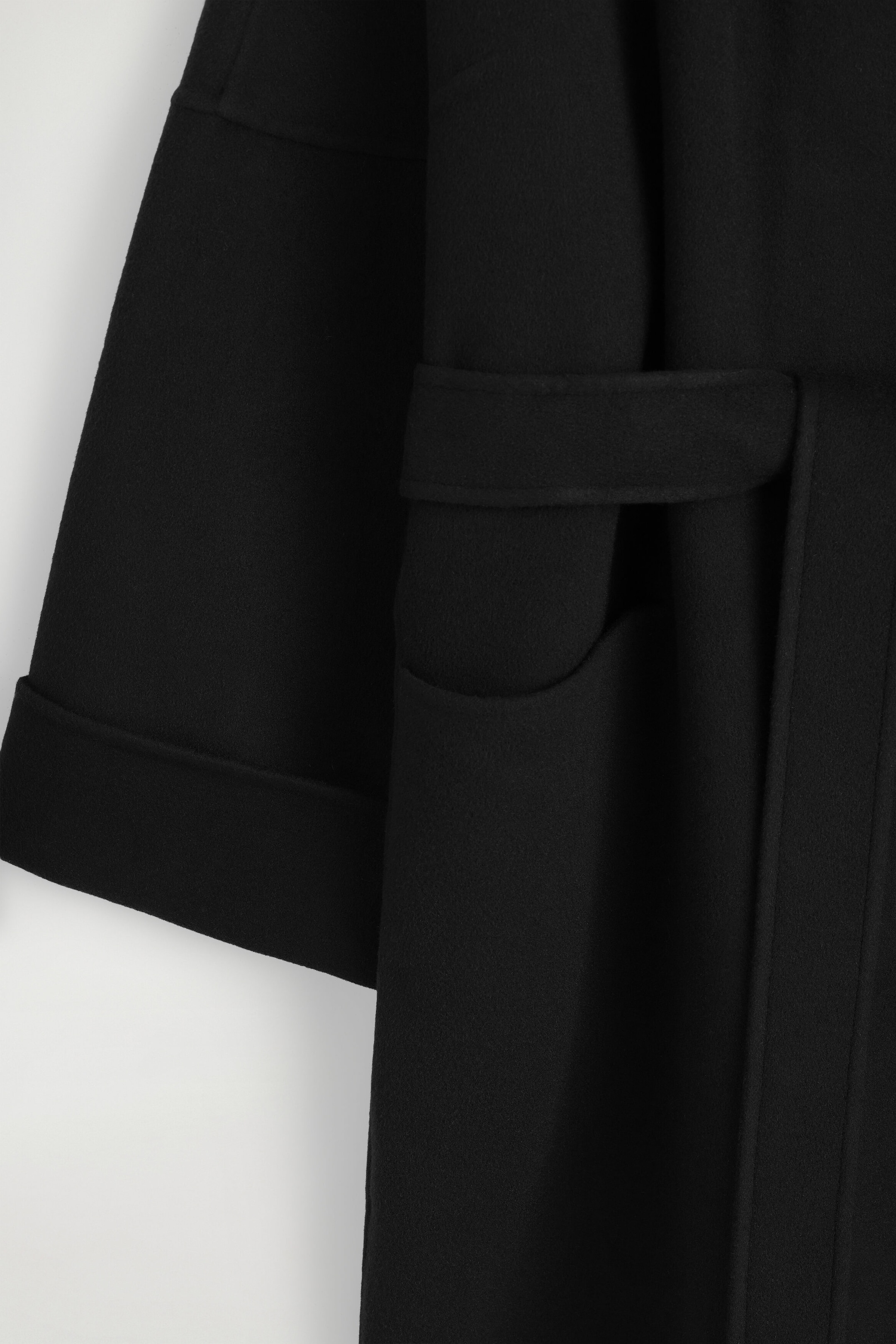 Ce gros plan du manteau châle oversize en laine double face noire montre une manche large avec un large revers replié en profondeur, une ceinture assortie dans le même tissu passant dans une boucle de ceinture, ainsi qu'une poche plaquée incurvée. Ce gros plan du manteau châle oversize en laine double face noire montre une manche large avec un large revers replié en profondeur, une ceinture assortie dans le même tissu passant dans une boucle de ceinture, ainsi qu'une poche plaquée incurvée.