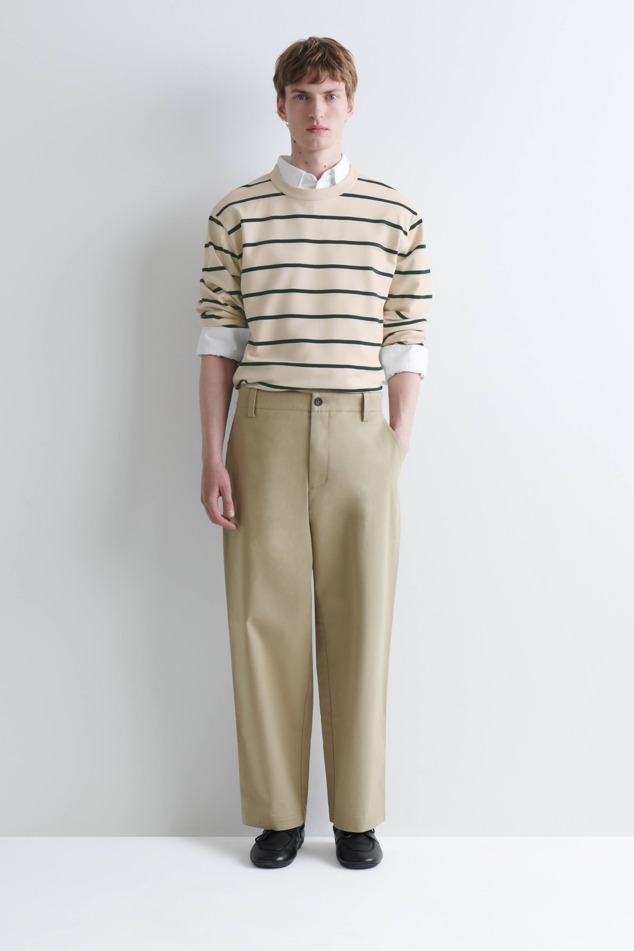 View larger image: Cotton twill relaxed-fit straight-leg trousers in a beige hue are being worn by the model, who is standing with one hand resting in a side pocket. The trousers feature a front button fastening and belt loops, and are paired with a striped crewneck sweatshirt and black shoes.