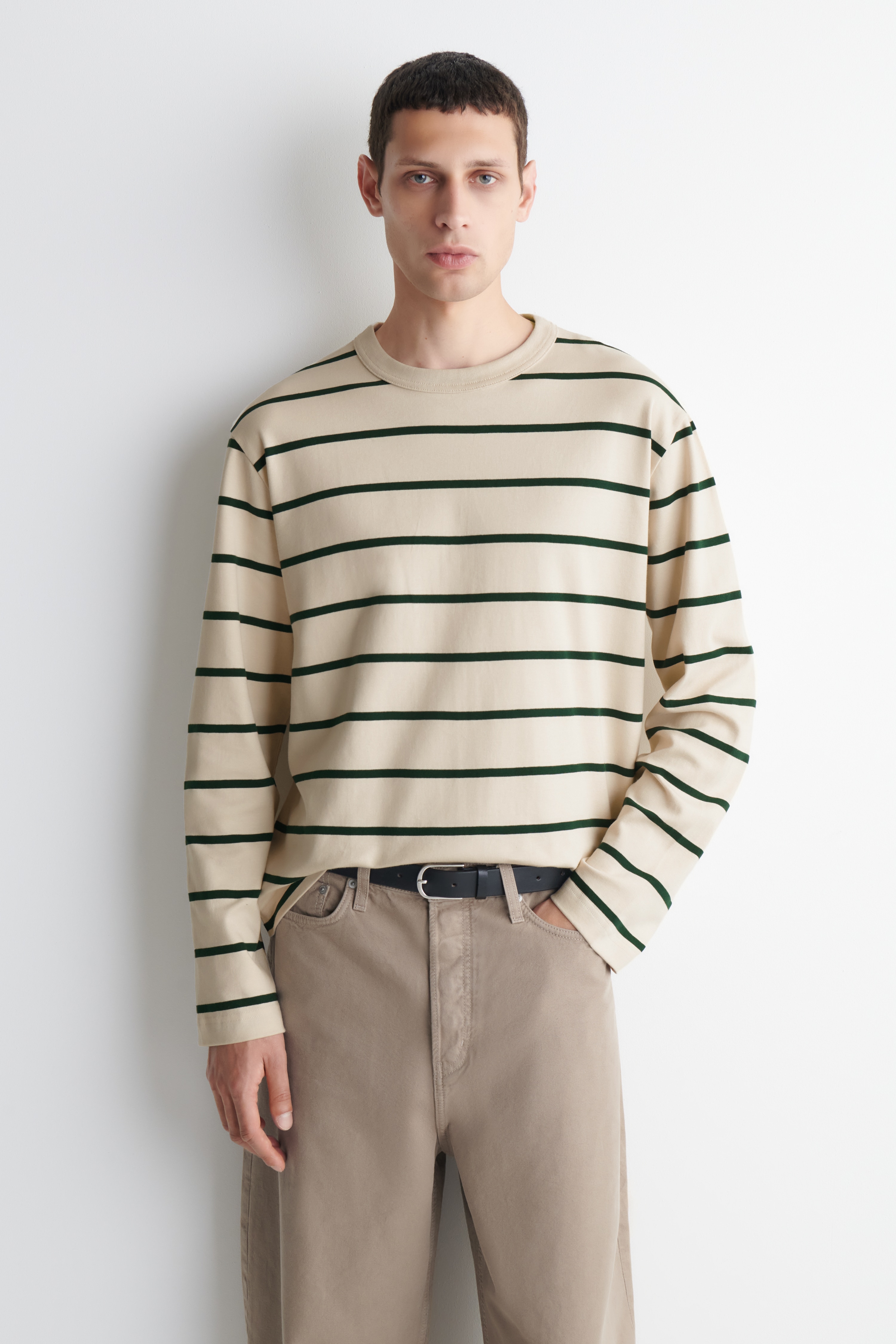 Uma camisola de manga comprida e decote redondo estÃ¡ virada para a frente, exibindo uma cor bege com riscas horizontais em verde escuro. A peÃ§a apresenta um corte descontraÃ­do com ombros descaÃ­dos, mangas compridas largas e uma bainha direita.